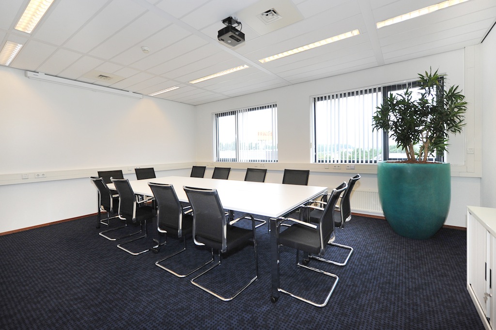 Vergaderruimte aan de Laan van ’s-Gravenmade met een lange tafel, zwarte stoelen, grote plantenbak en een projector aan het plafond.
