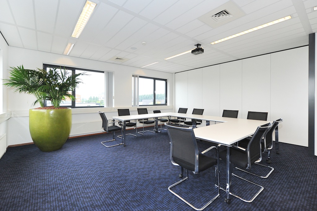 Moderne vergaderruimte aan de Laan van S-Gravenmade met een U-vormige opstelling van tafels, zwarte stoelen en een grote groene plantenbak.