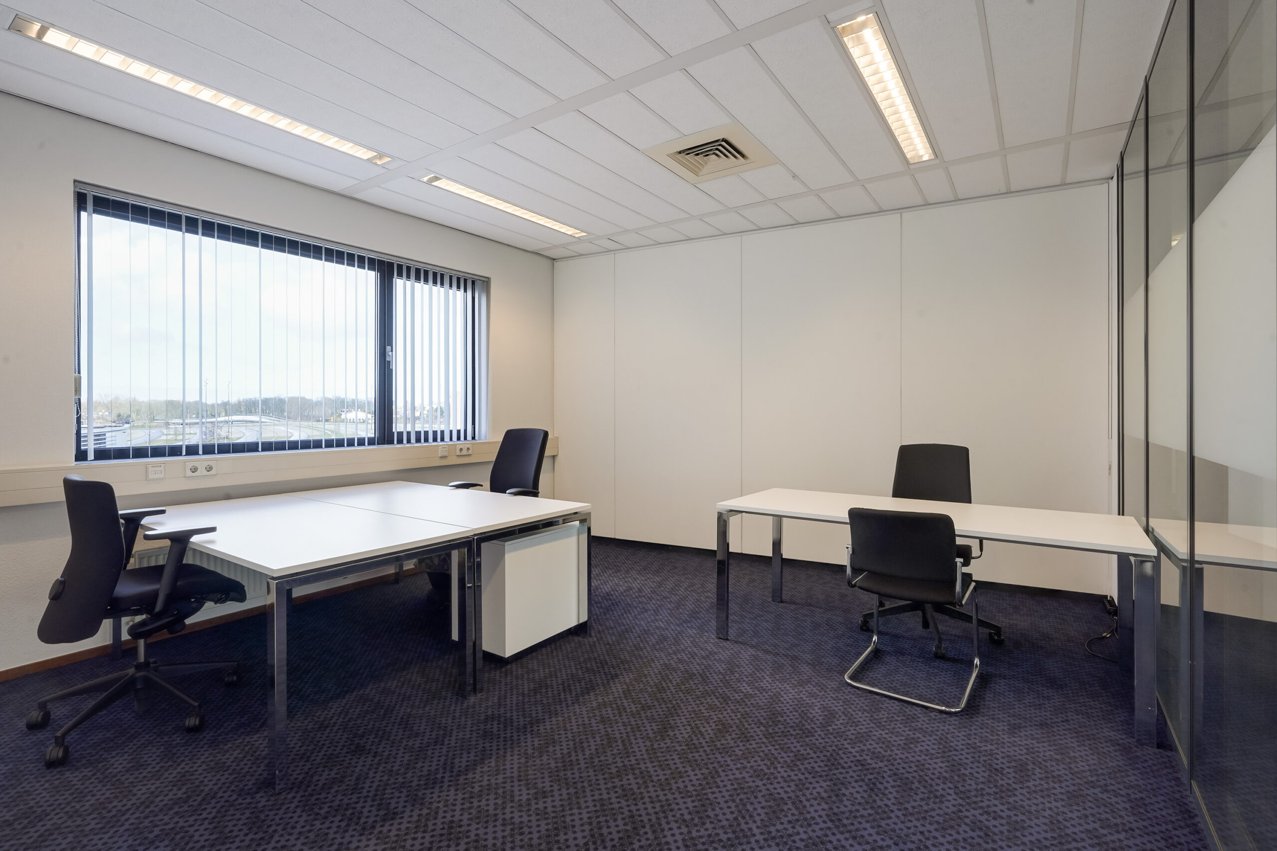 Moderne, lichte kantoorruimte aan de Laan van S-Gravenmade met drie bureaus, bureaustoelen en een groot raam met verticale lamellen.