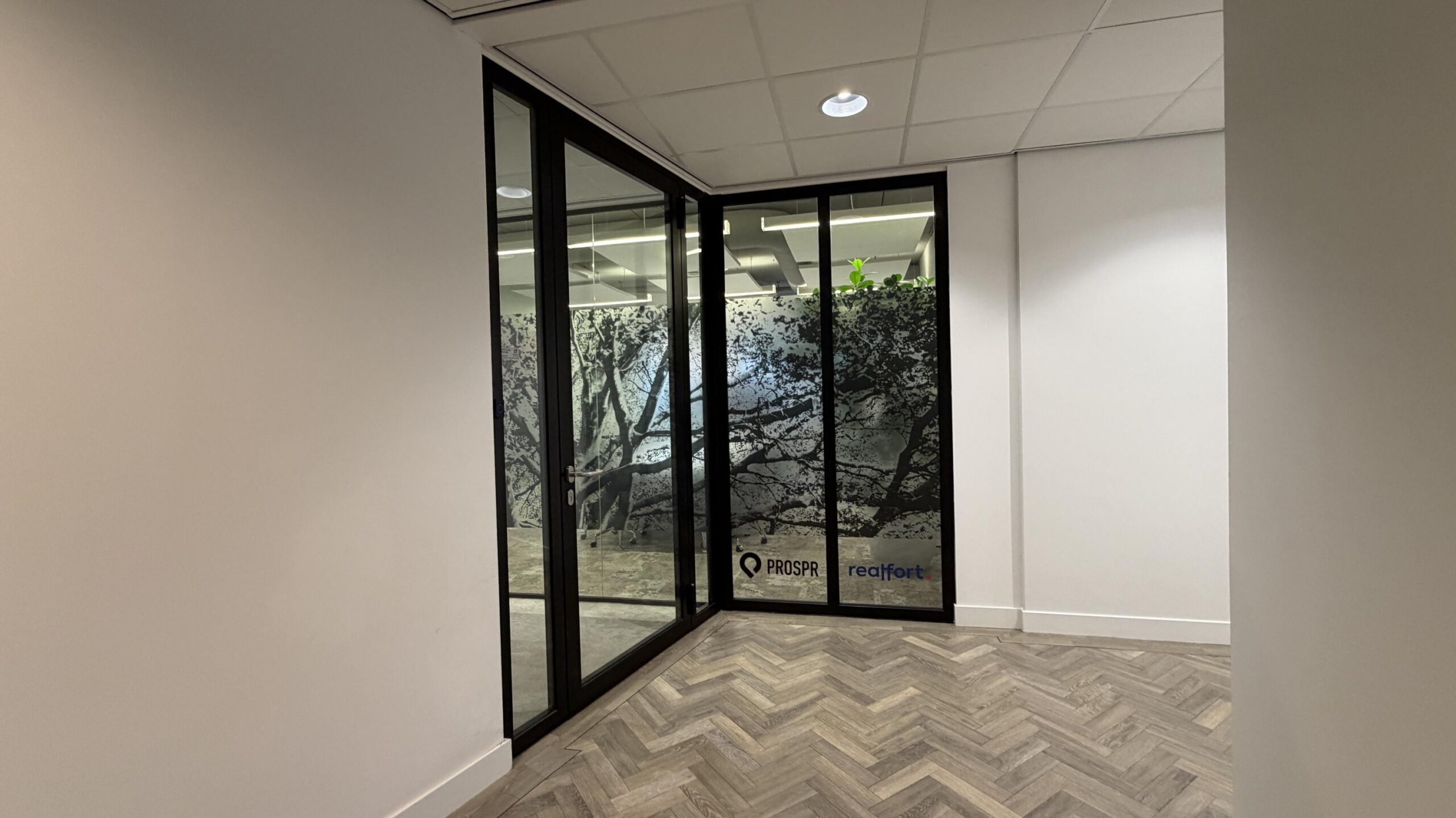 Ingang van een modern kantoor aan de Euclideslaan met glazen wanden en decoratieve boomprint, met logo’s van PROSPR en realfort.