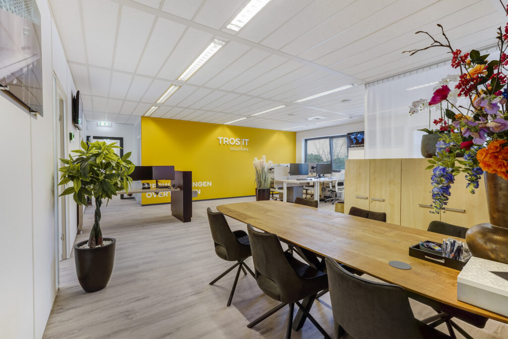 Moderne kantooruimte van Tros IT Solutions met een gele accentmuur, bureaus, vergadertafel en kleurrijke bloemenvaas.
