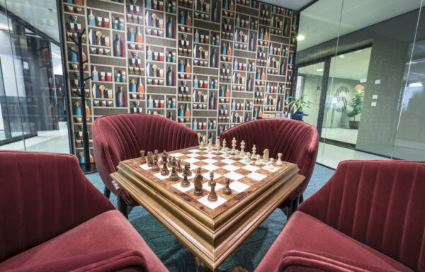 Een schaakbord met opgestelde stukken staat centraal tussen drie rode stoelen in een modern ingerichte kamer.