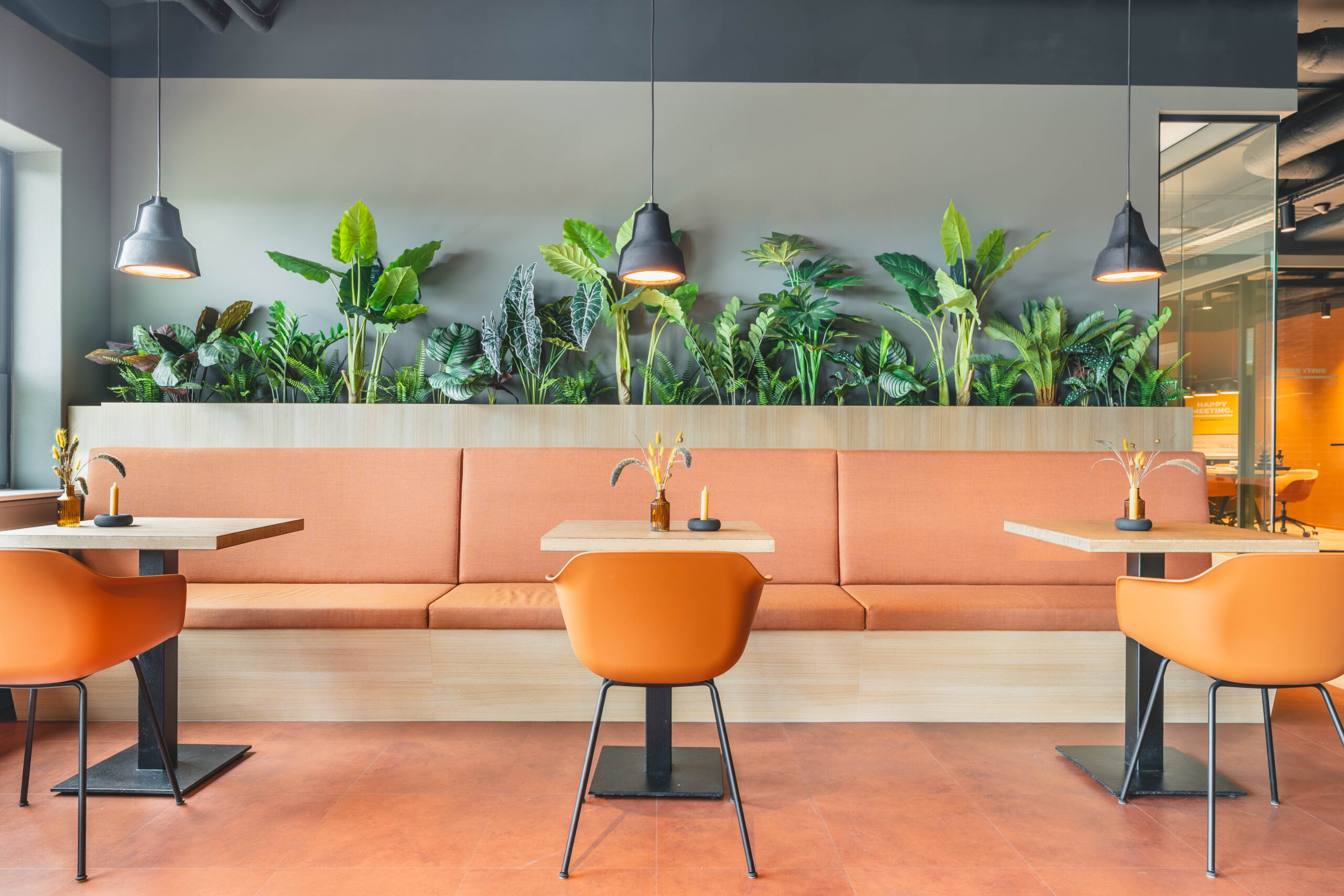 Een moderne café-interieur met oranje stoelen en bankjes, houten tafels en groene planten op de achtergrond.