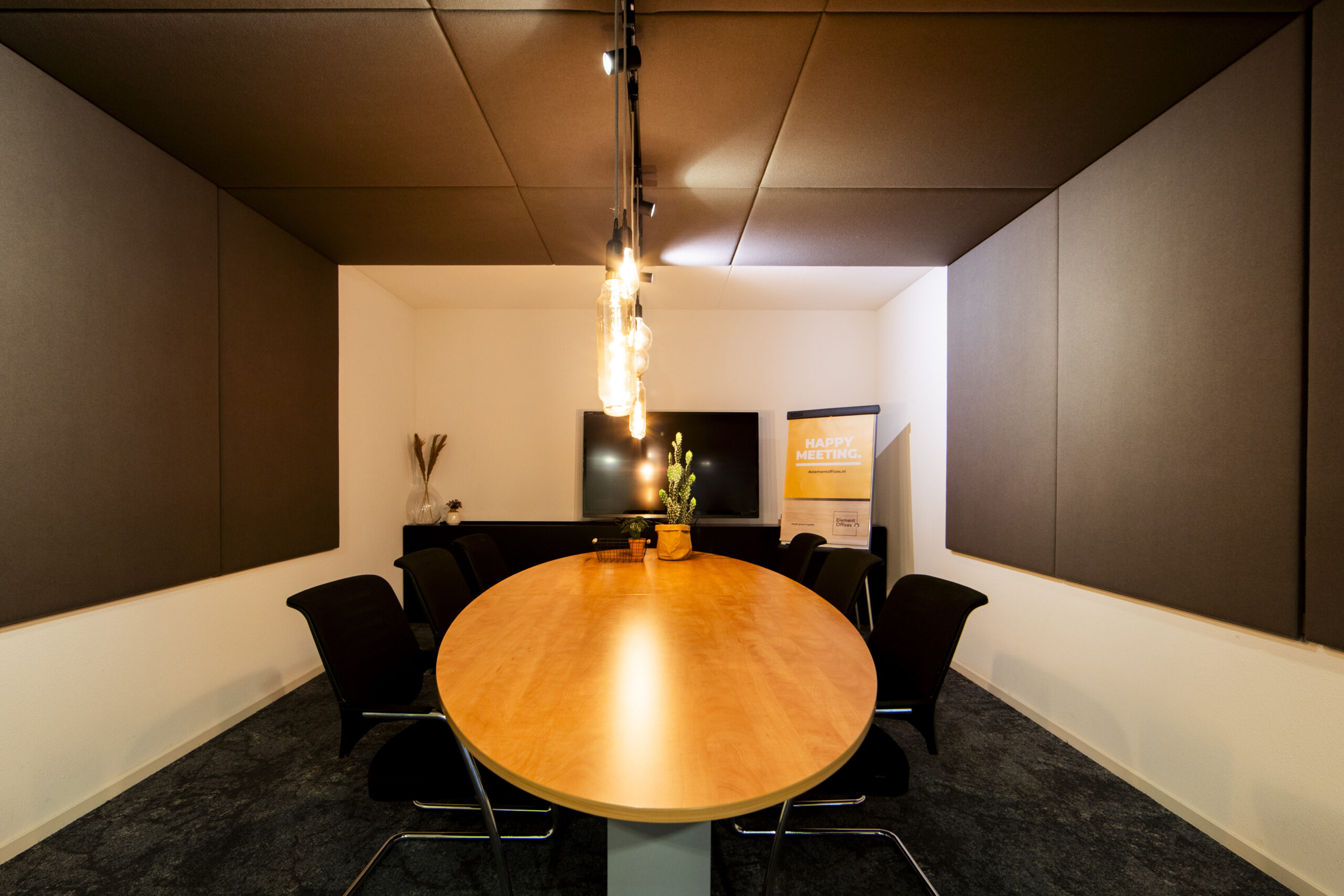 Vergaderruimte met een ovale houten tafel, stoelen, een beeldscherm en een posters met de tekst "Happy Meeting".