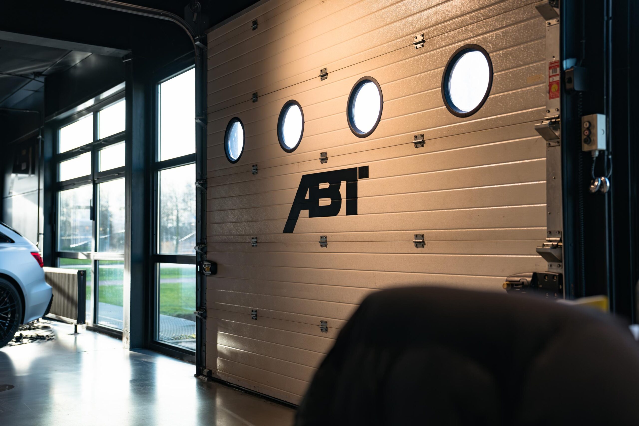 Garage met een grote witte roldeur met ABT-logo en ronde ramen, deels geopend naast een geparkeerde auto.