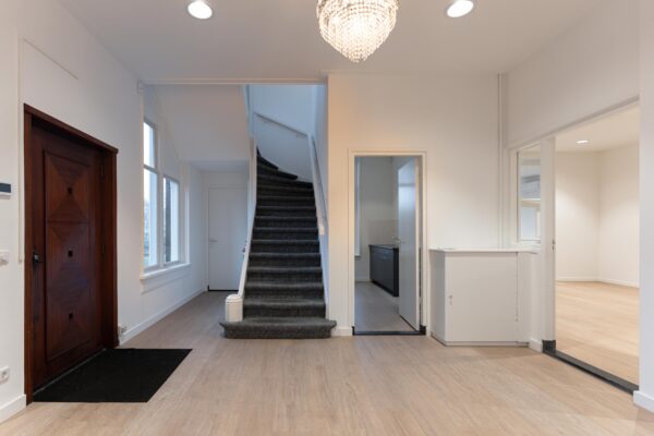 Hal met houten voordeur, trap met grijze vloerbedekking, en doorgangen naar keuken en andere kamers in een woning aan de Biltstraat.