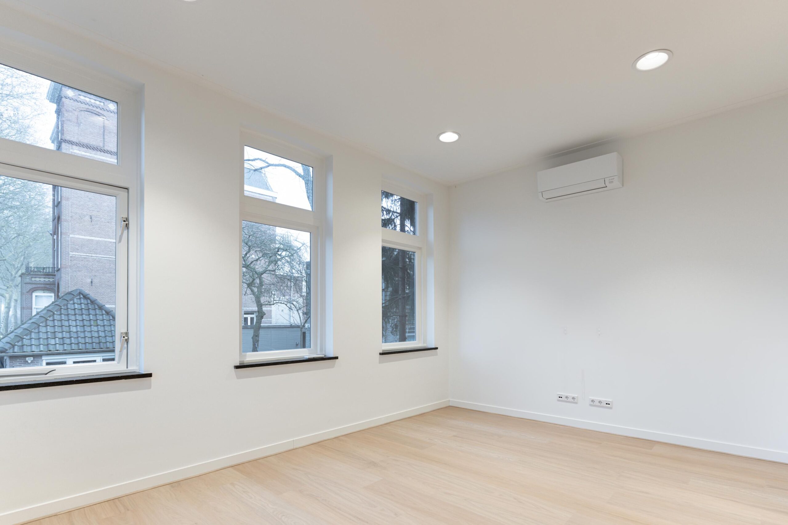 Licht, leeg vertrek met houten vloer, grote ramen en een airco aan de muur in een pand aan de Biltstraat.