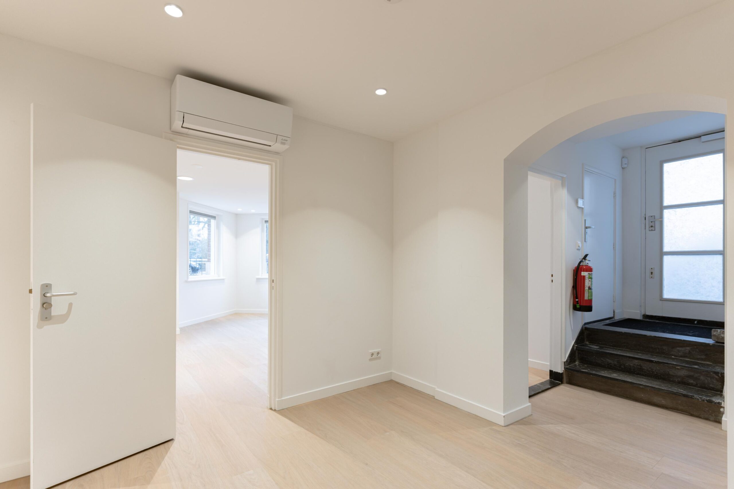 Lichte hal met een open deur naar een kamer met ramen, een airco aan het plafond en een trap bij de voordeur.