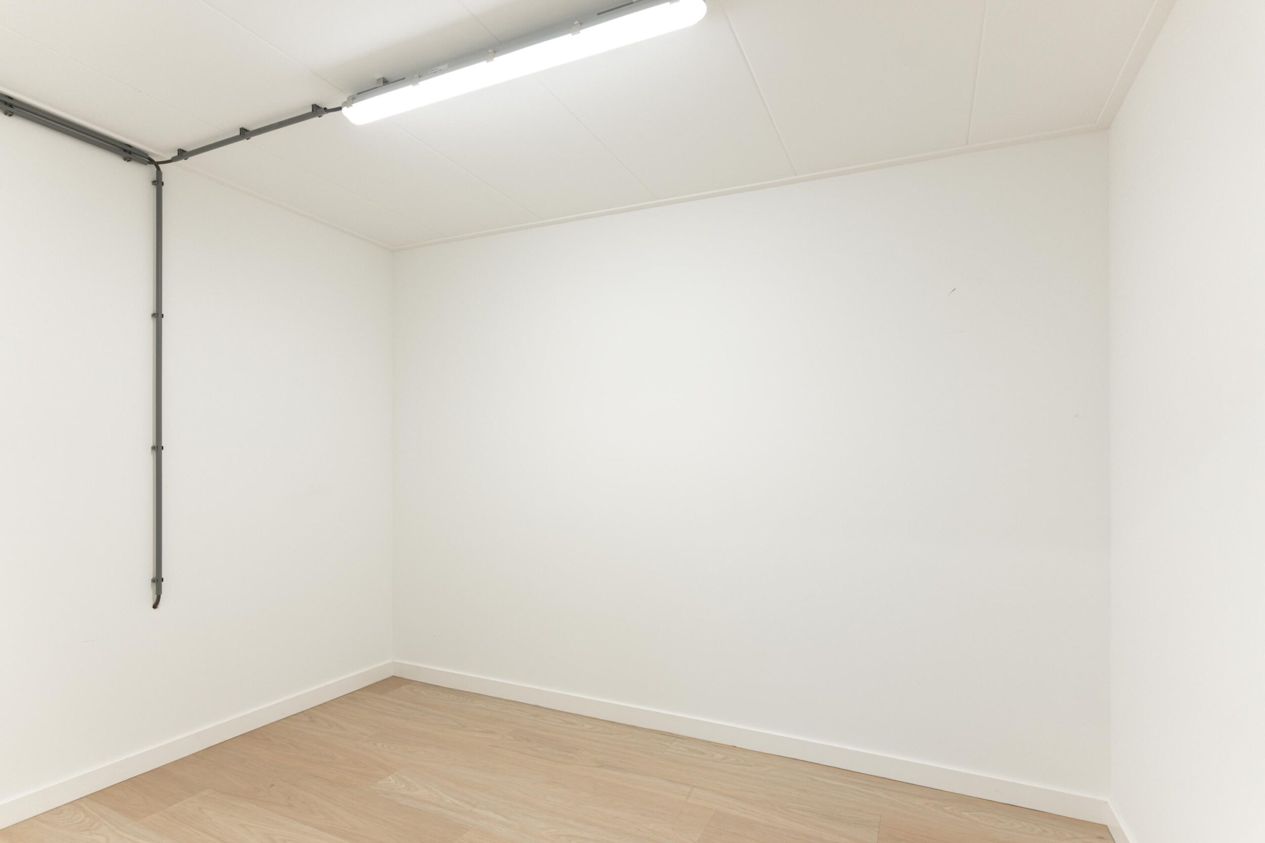Leeg, witgeschilderd kamertje met een houten vloer en zichtbare leiding langs het plafond aan de Biltstraat.