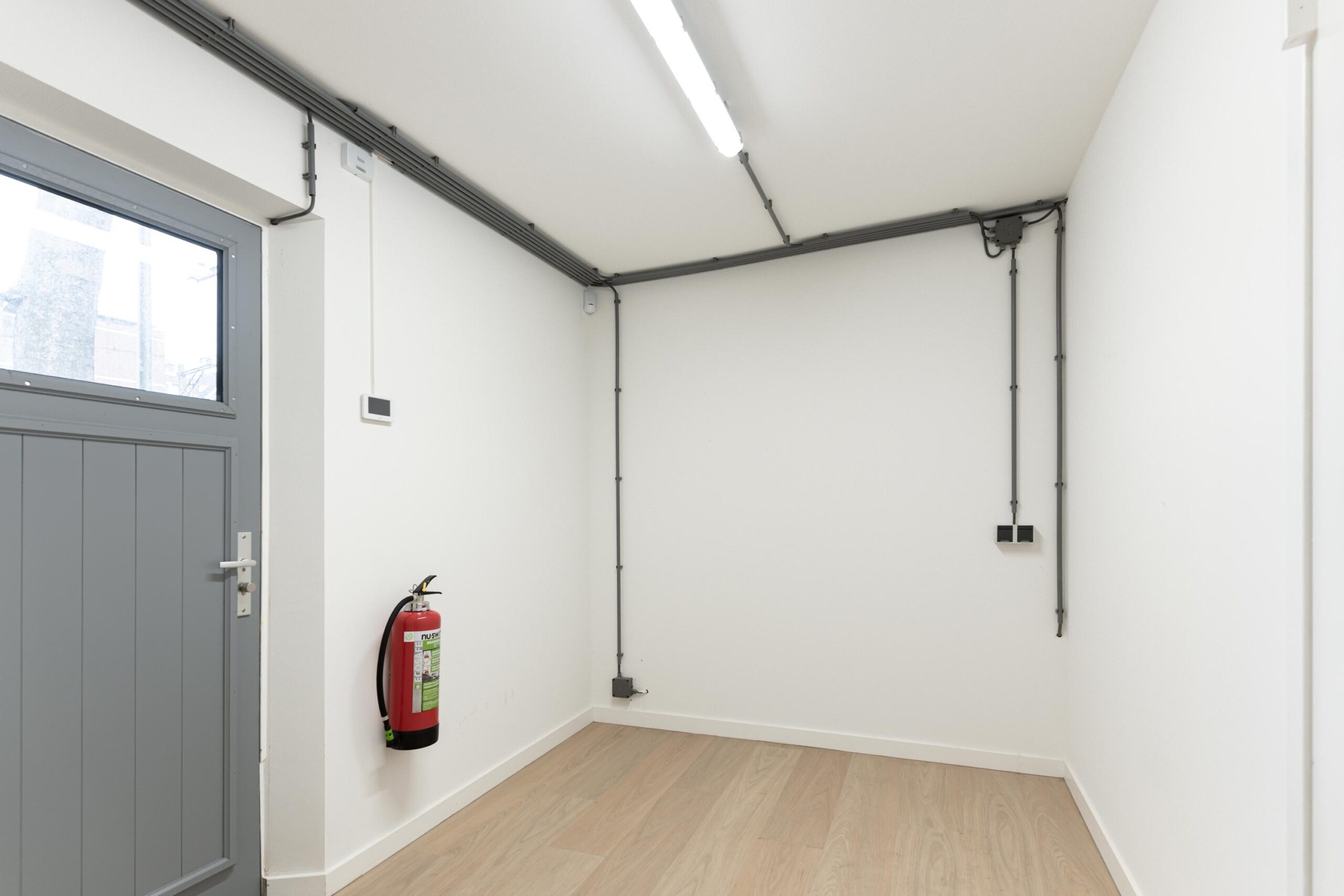 Lege witte kamer met houten vloer, grijze deur, brandblusser en zichtbare elektrische bekabeling aan het plafond.