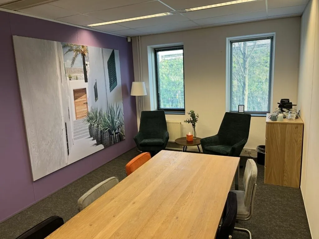 Vergaderruimte met houten tafel, kleurrijke stoelen, twee groene fauteuils en een grote wanddecoratie met afbeelding van een zonnige steeg.