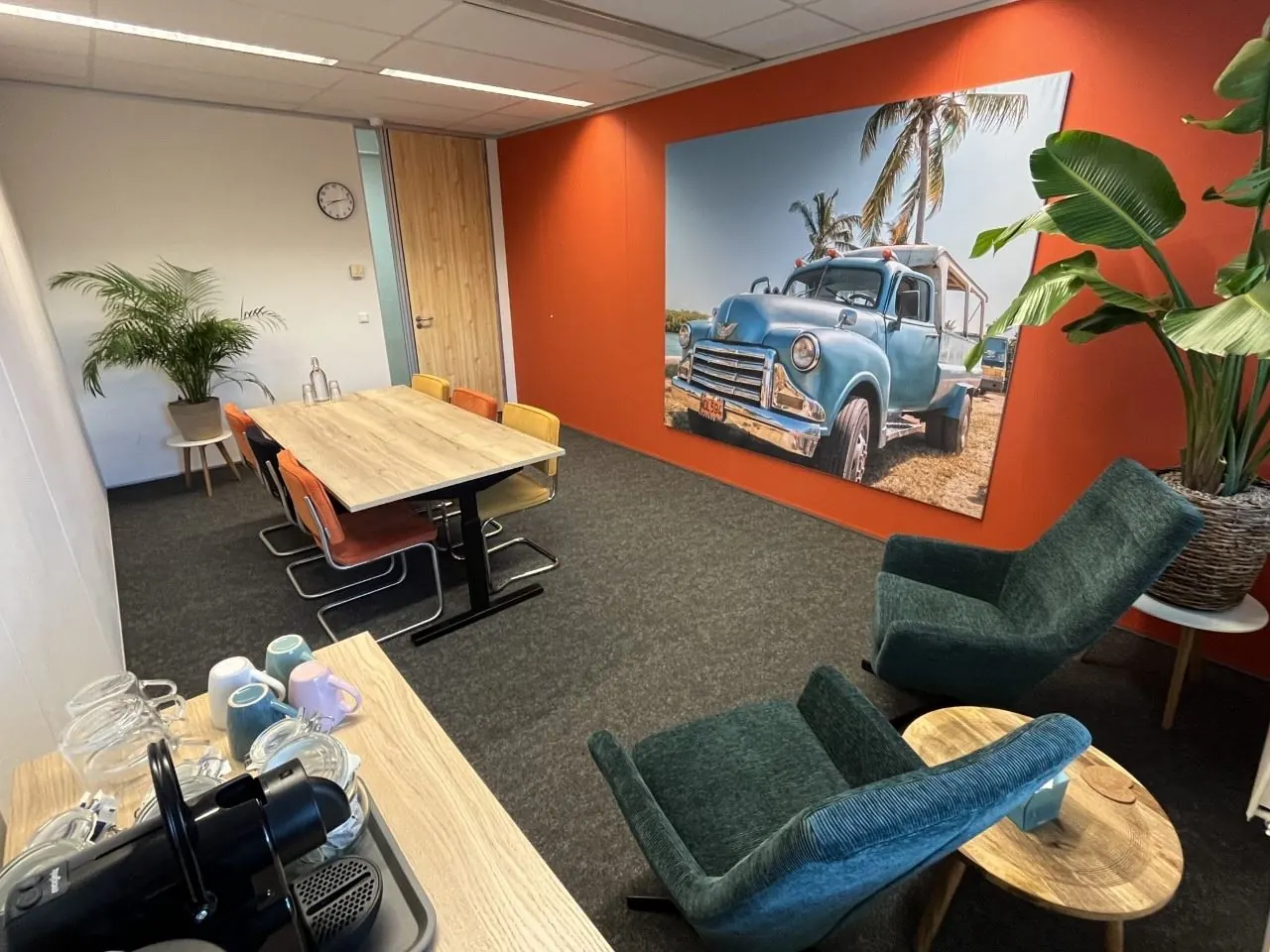Moderne vergaderruimte met koffiehoek, kleurrijke stoelen, grote plant en een opvallende wanddecoratie van een blauwe oldtimer met palmbomen (titel: 269_2160).