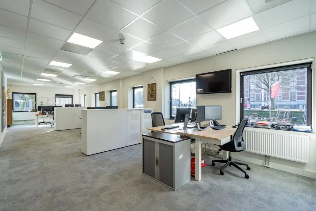 Modern, licht kantoor met meerdere bureaus, computers en grote ramen met uitzicht op een straat.