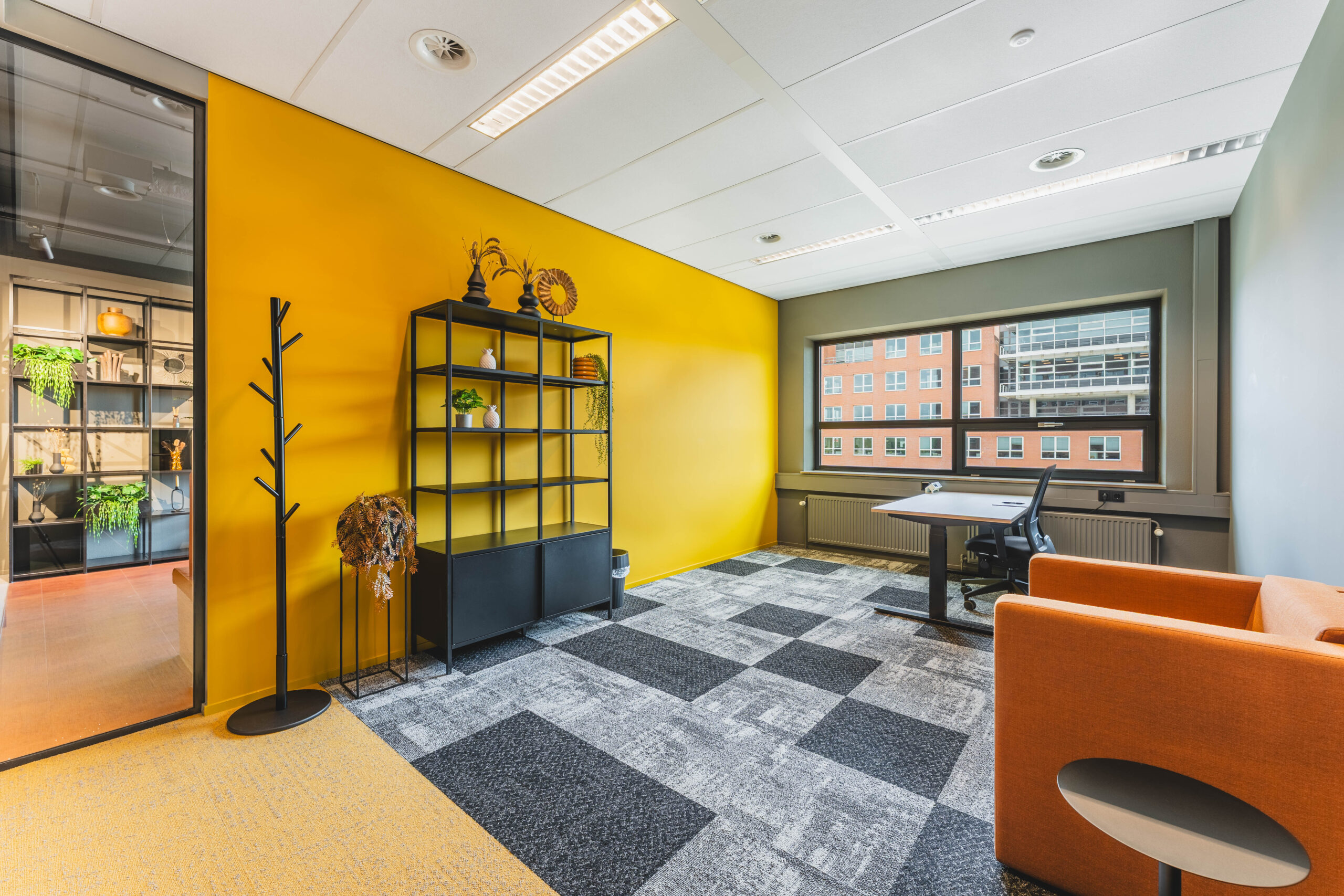 Een moderne kantoorruimte met een gele muur, een zwart open rek en een oranje stoel bij het raam met uitzicht op een bakstenen gebouw.