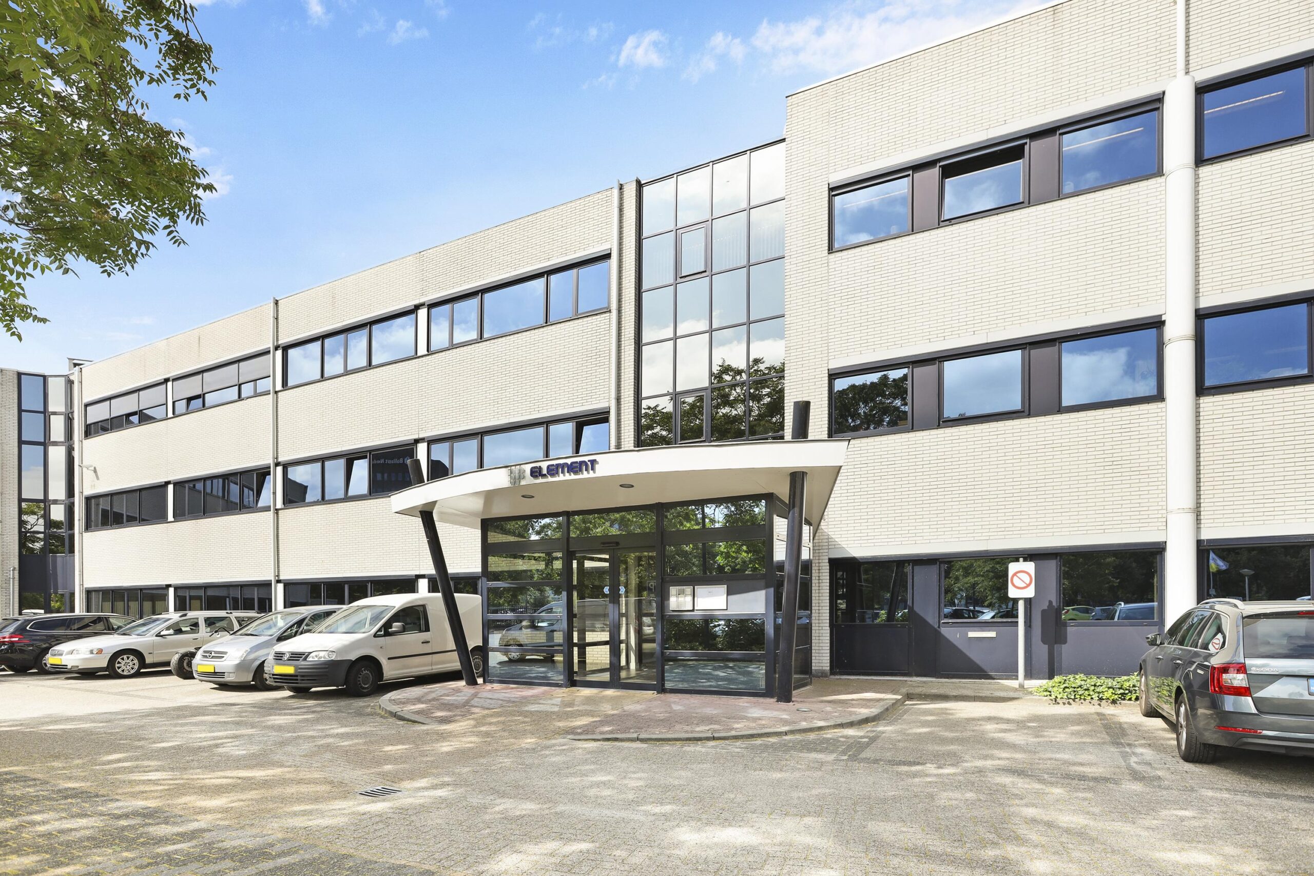 Voorzijde van een modern kantoorgebouw met glazen entree en meerdere geparkeerde auto's ervoor.