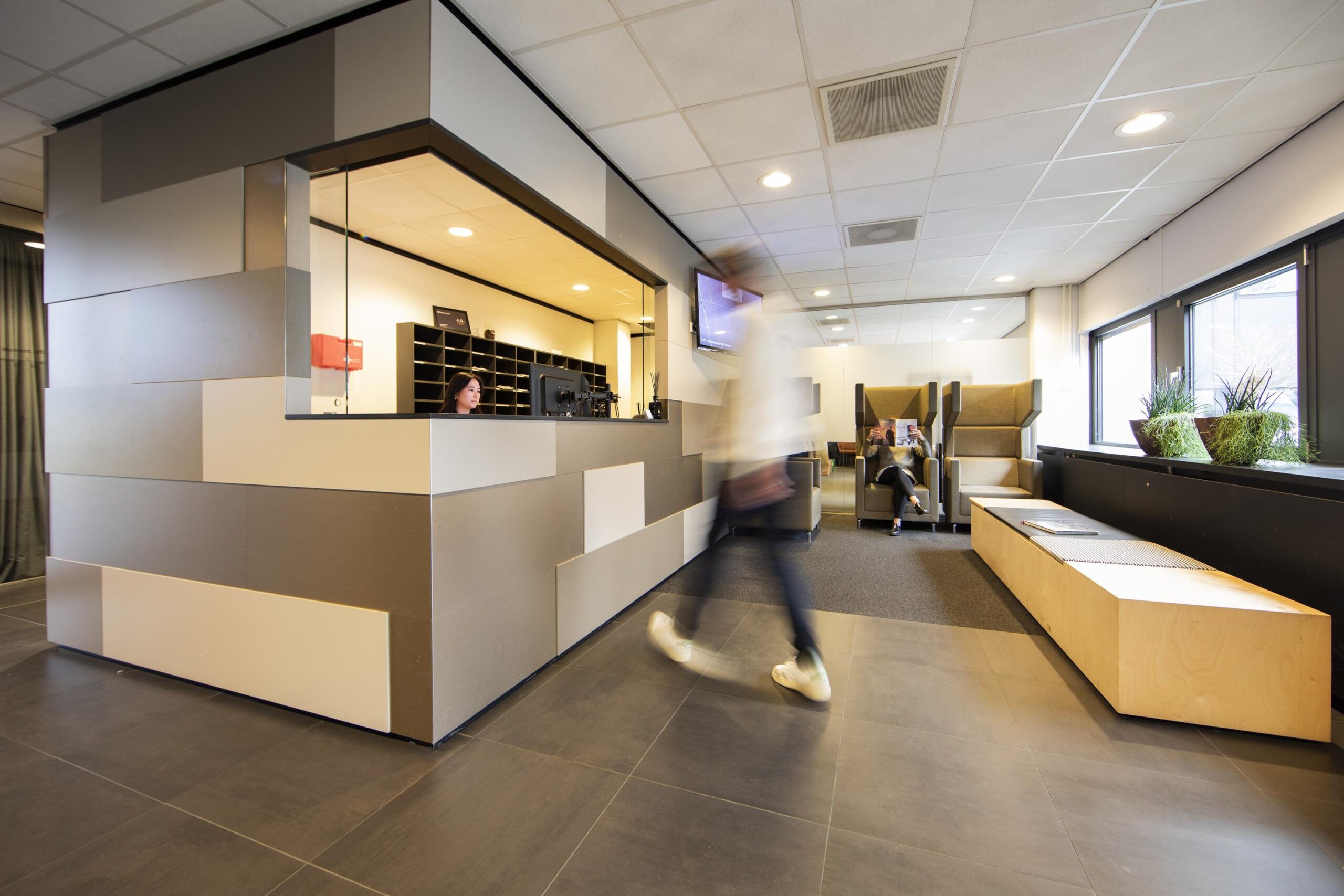 Een moderne receptie met een medewerker achter een balie en een persoon die voorbij loopt, naast een wachtruimte met zithoek en planten.