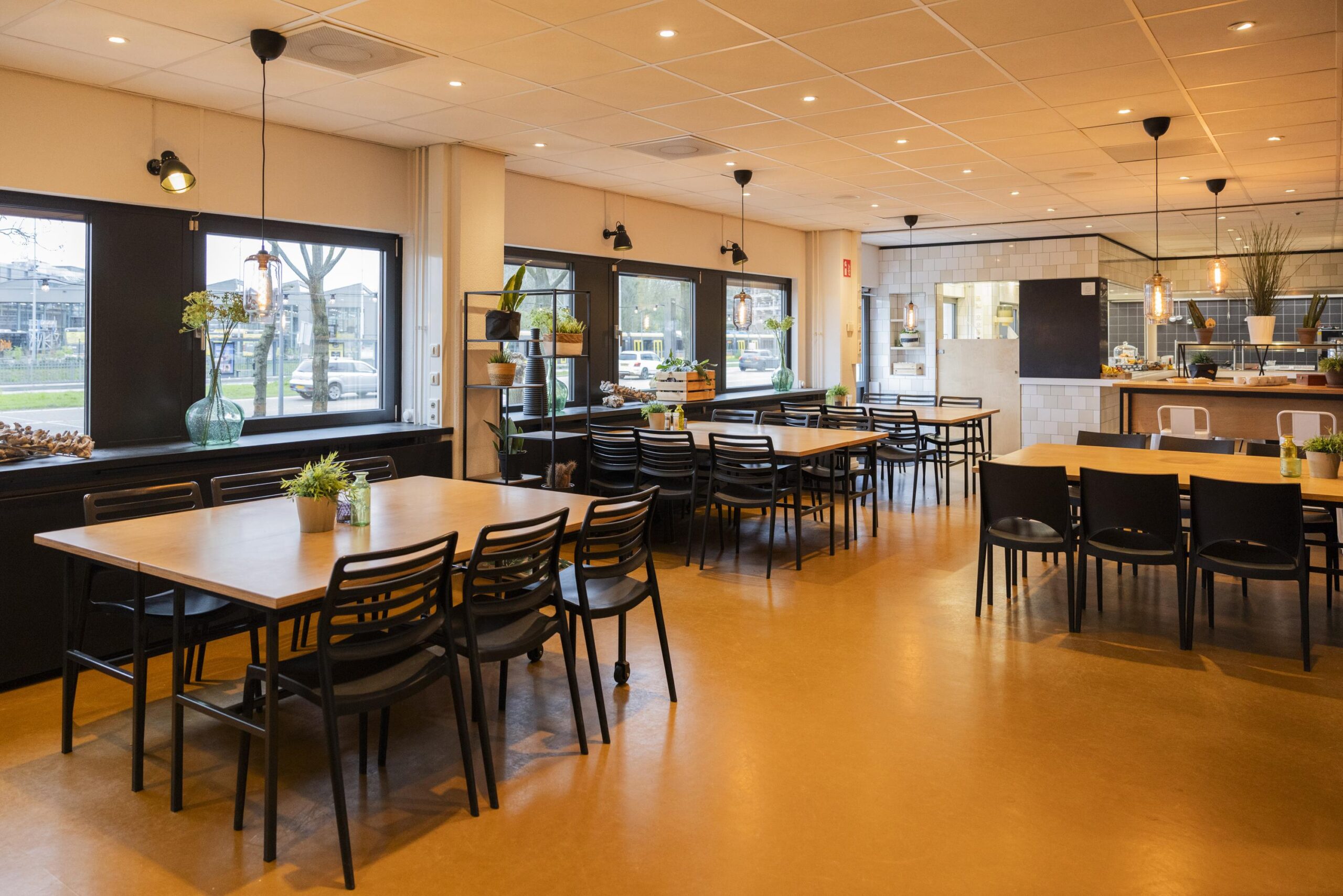Een ruime, moderne kantine met houten tafels, zwarte stoelen en decoratieve planten.