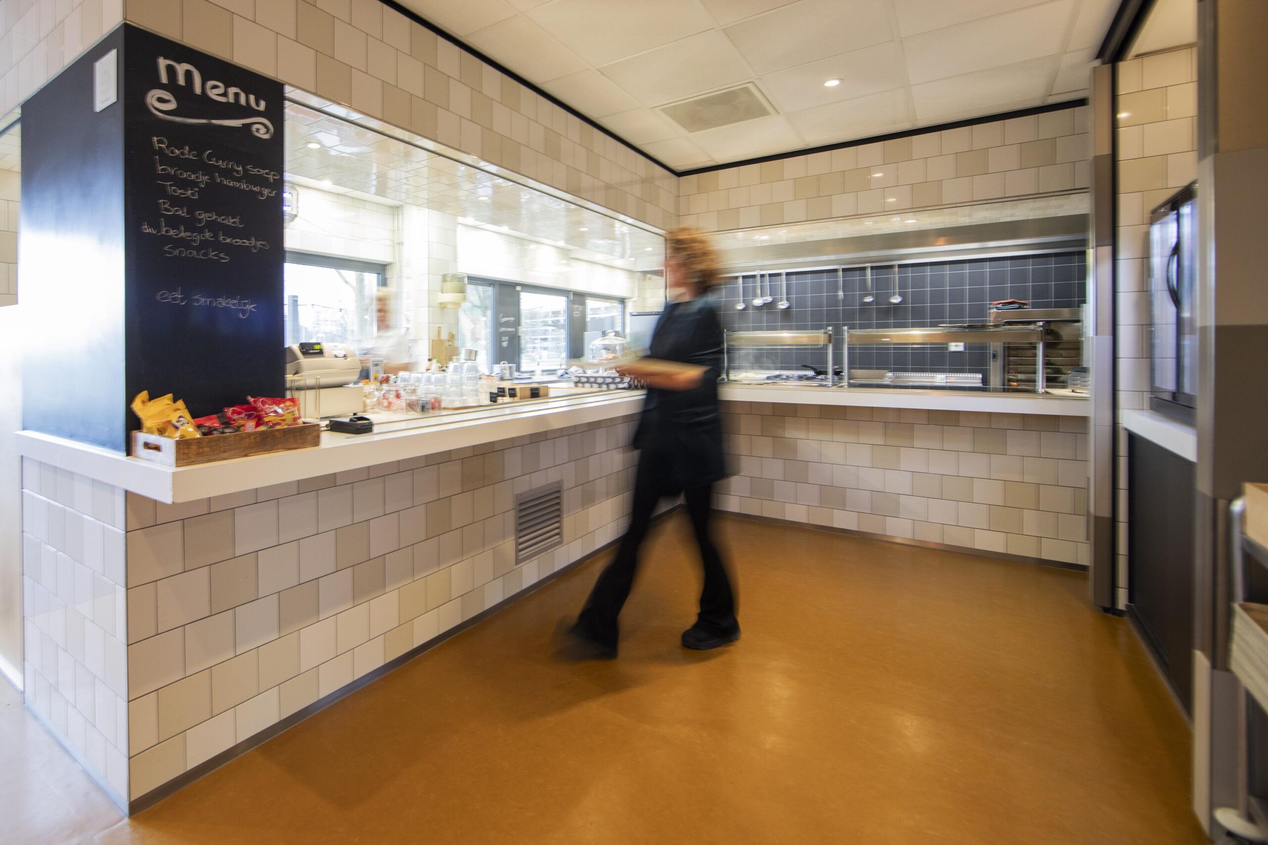 Een vrouw loopt langs een balie in een moderne kantine met een menubord op de achtergrond.