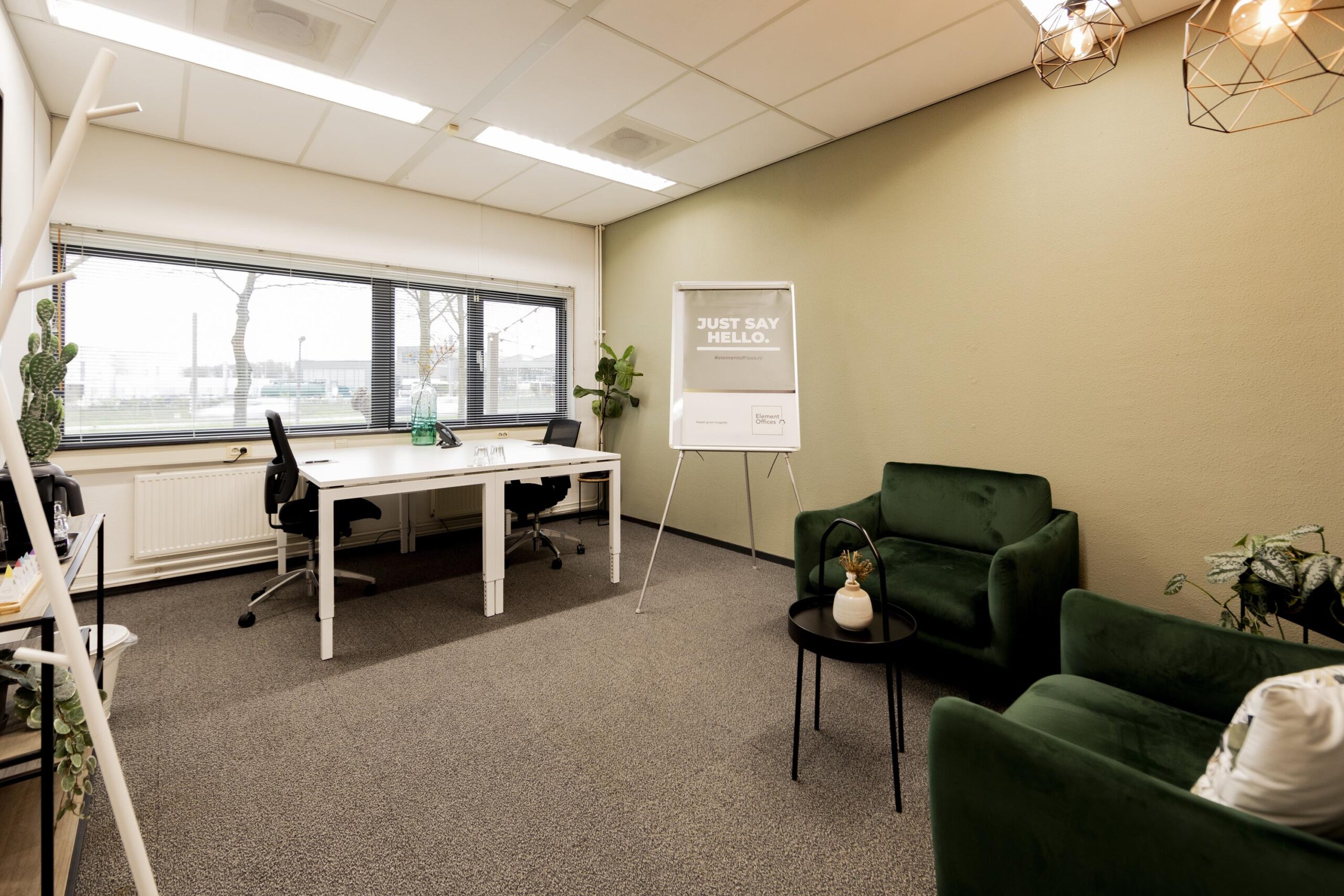 Een modern ingerichte kantoorruimte met een groot raam, twee groene fauteuils, een bureau en een flip-over met de tekst "Just Say Hello."