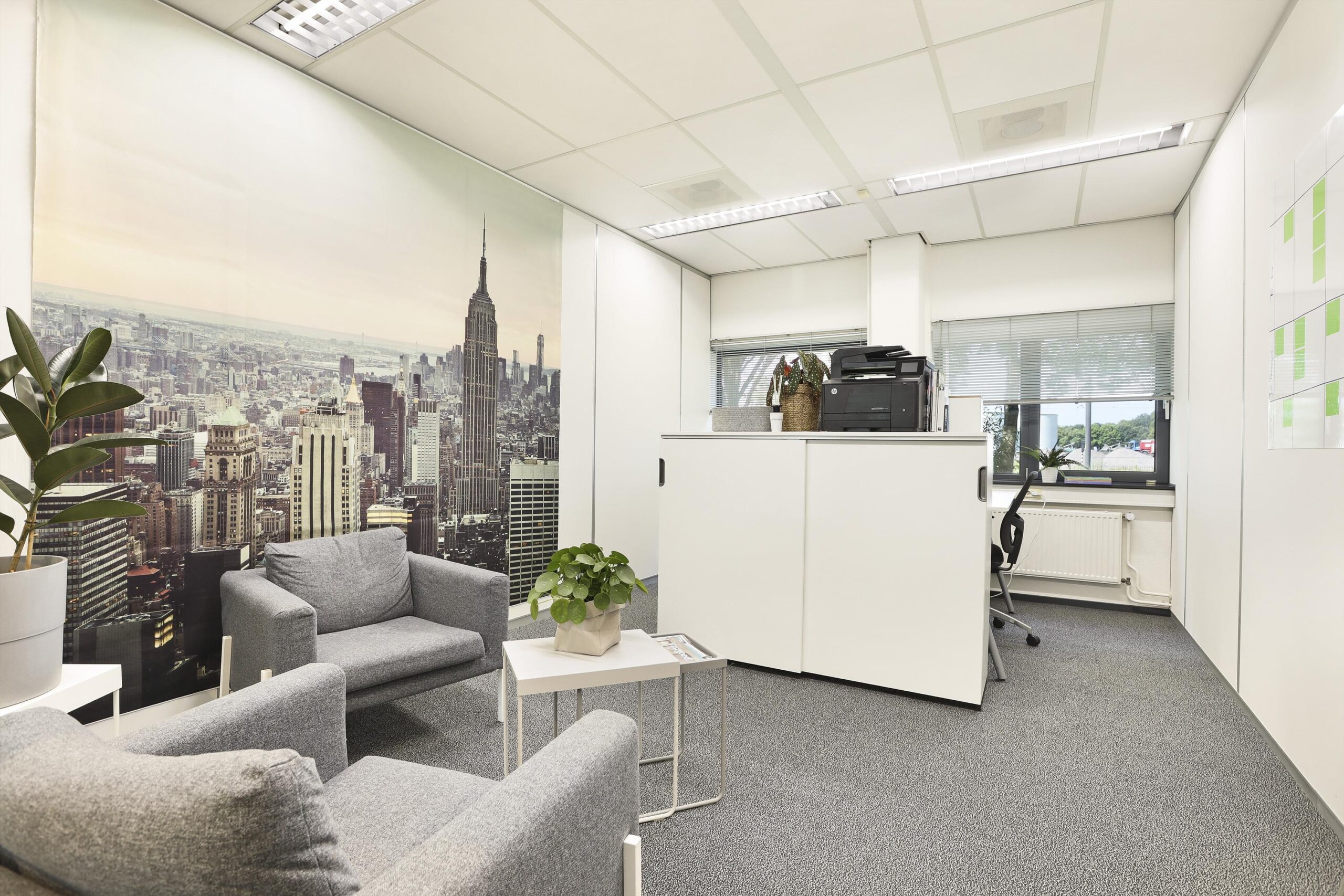 Een moderne kantoorruimte met grijze stoelen, een tafel met planten en een fotobeeld van een skyline op de achtergrond.