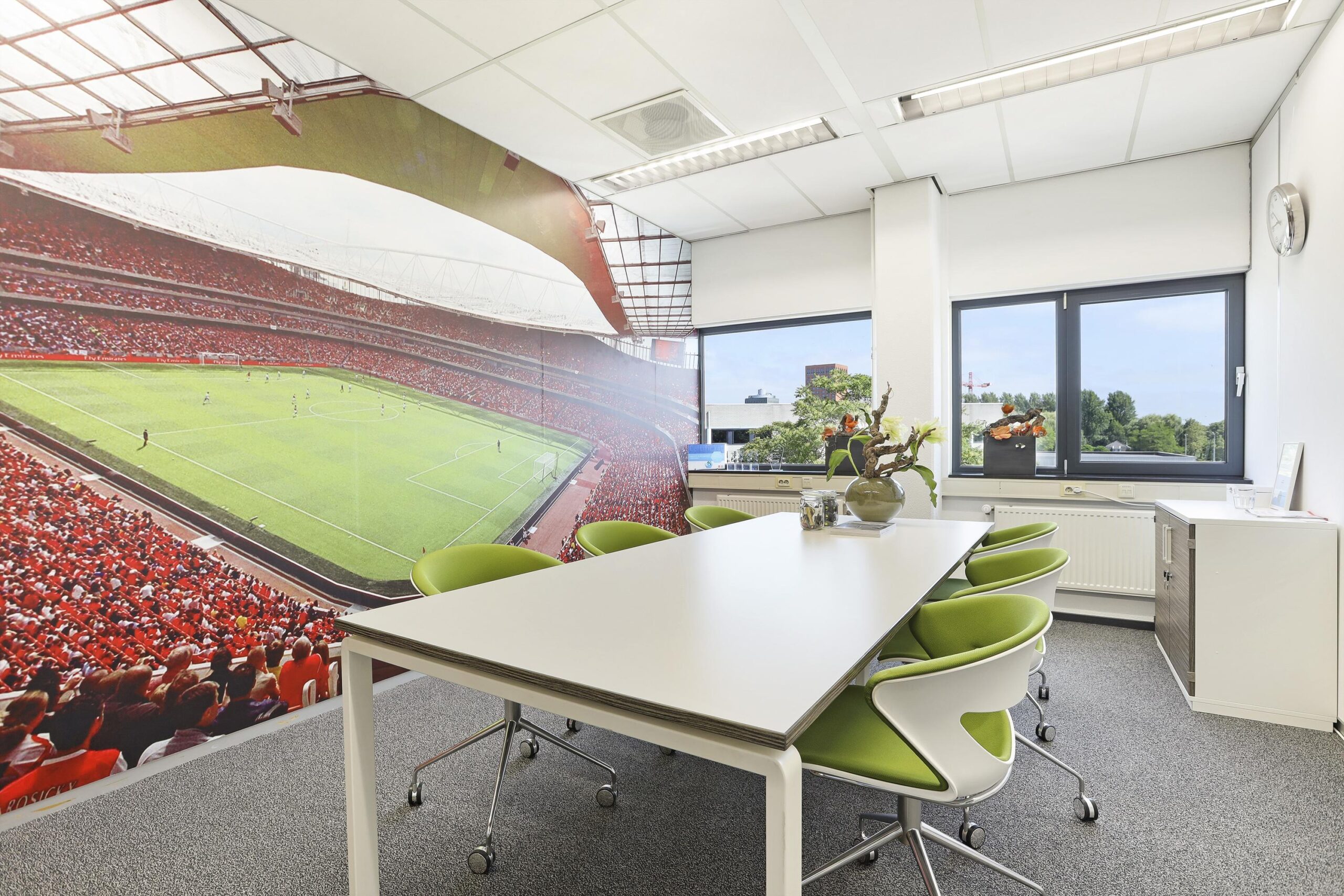 Een vergaderruimte met een grote muurschildering van een voetbalstadion en moderne meubels met groene stoelen.