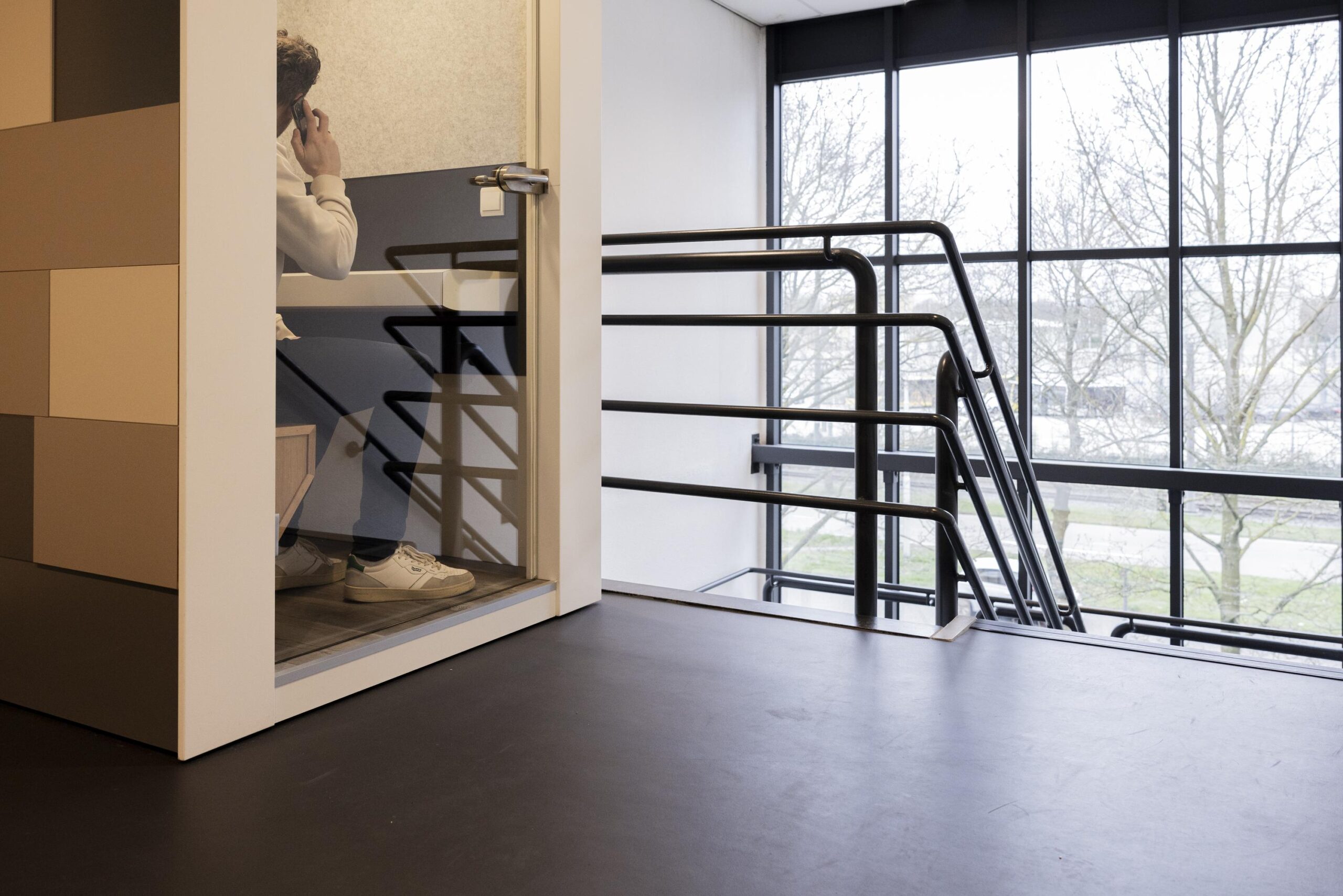 Een persoon zit in een glazen cabine en is aan het bellen, met een trap en grote ramen op de achtergrond.