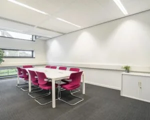 Vergaderruimte met witte tafel, acht roze stoelen en grote ramen met uitzicht op groen.