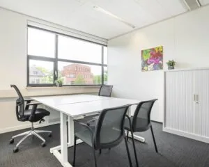 Moderne, lichte kantoorruimte met een vergadertafel, vier stoelen, een raam met uitzicht en een kleurrijk schilderij aan de muur.