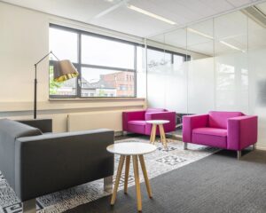 Moderne wachtruimte met grijze en felroze fauteuils, ronde bijzettafels en grote ramen.