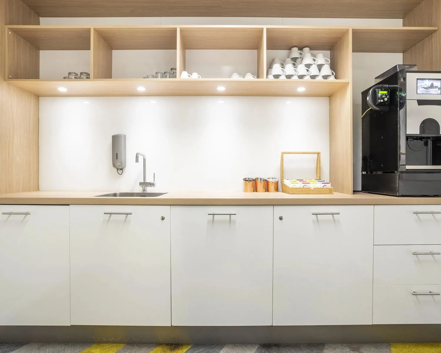 Moderne kantoorkeuken met een koffiemachine, spoelbak en open houten kasten met kopjes en glazen.