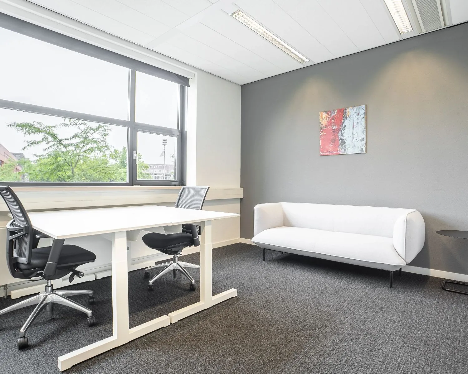 Modern ingericht kantoor met een wit bureau, twee zwarte bureaustoelen, een witte bank en een schilderij aan de grijze muur.