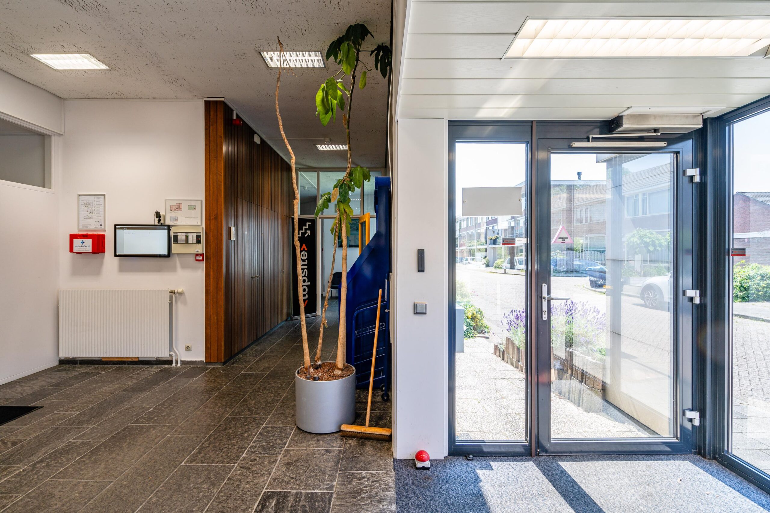 Ingang van een gebouw aan de Nicolaas Pieckstraat met deuren naar buiten en een ruime, lichte hal met een plant en schoonmaakgereedschap.