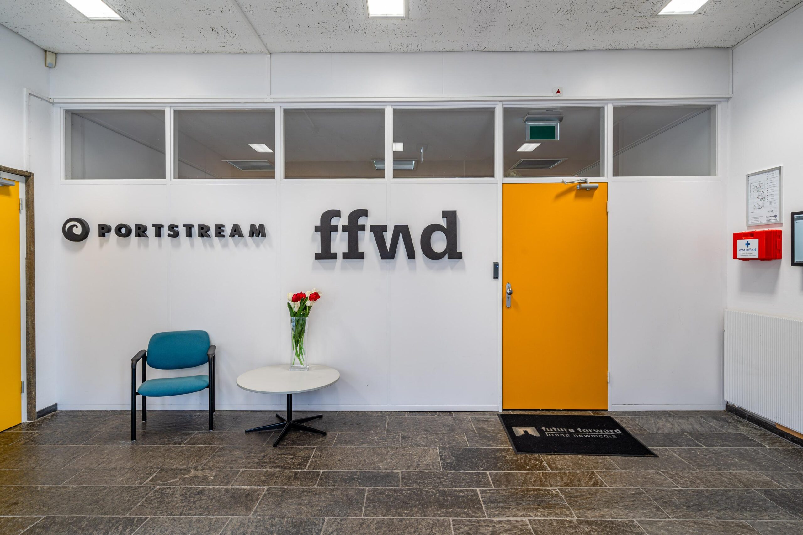 Entree met logo's van Portstream en ffvd, een stoel, een tafeltje met tulpen en een gele deur.