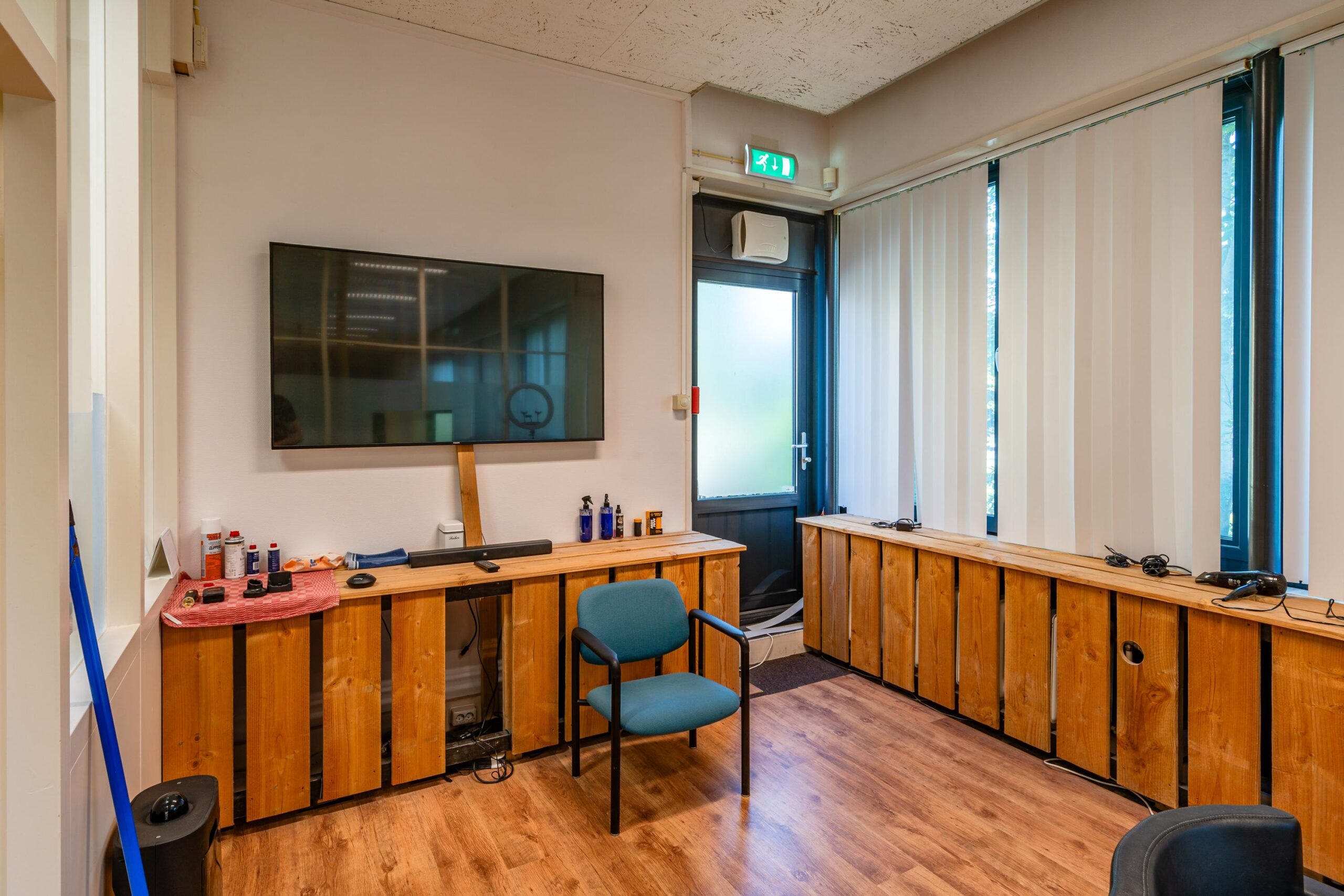 Een kantoorruimte met een houten vloer, een grote televisie aan de muur, een stoel en diverse apparatuur op een kast met houten panelen.