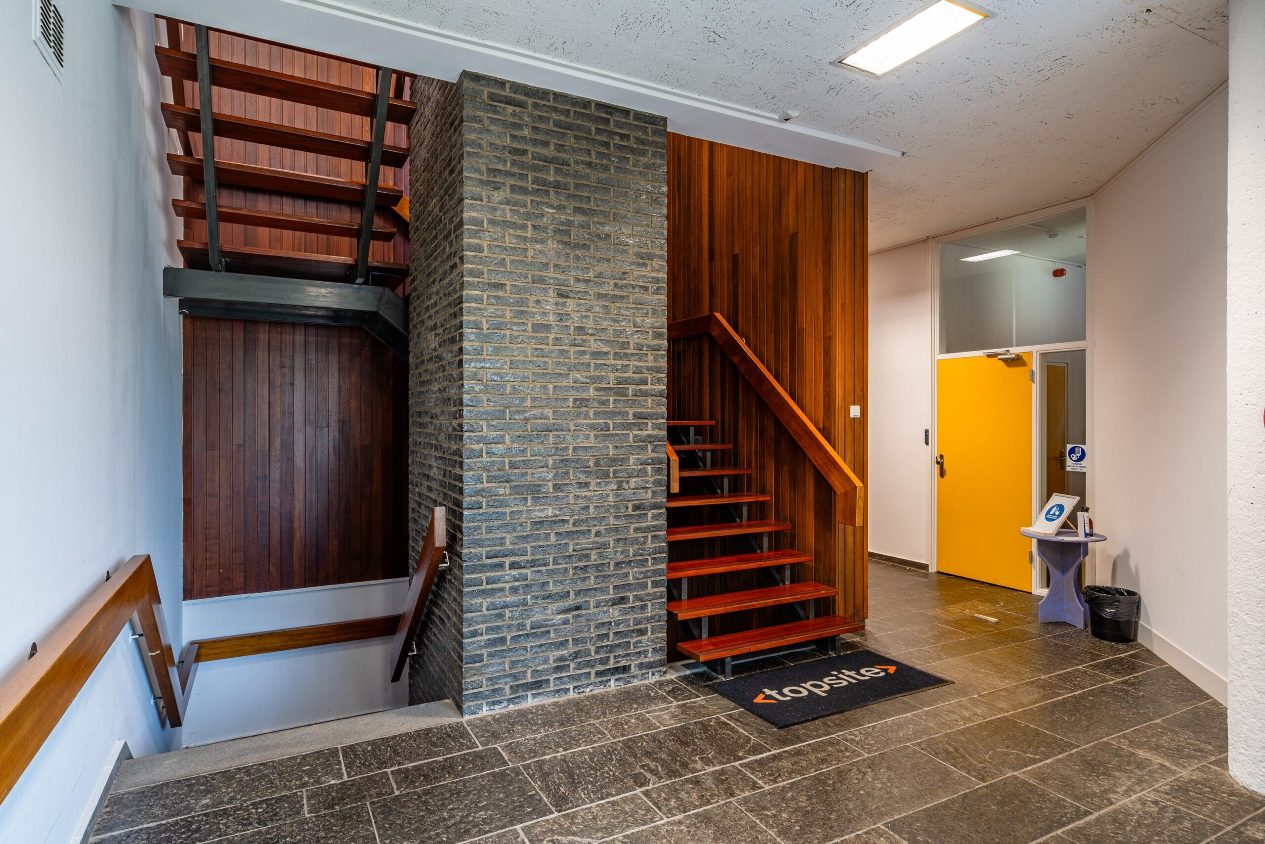 Een moderne entree met houten trap, bakstenen muur en felgele deur in de Nicolaas Pieckstraat.