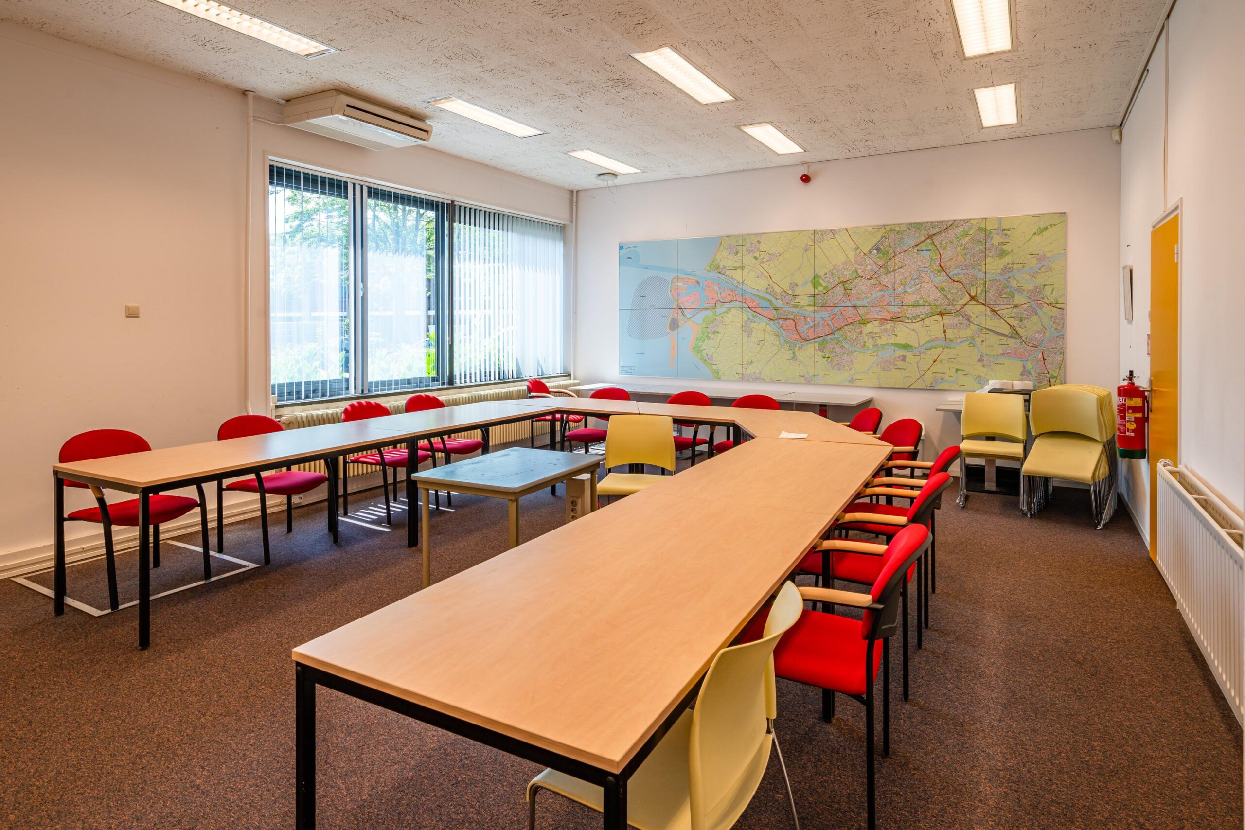 Een vergaderruimte met een U-vormige tafelopstelling, rode stoelen en een grote stadskaart aan de muur.