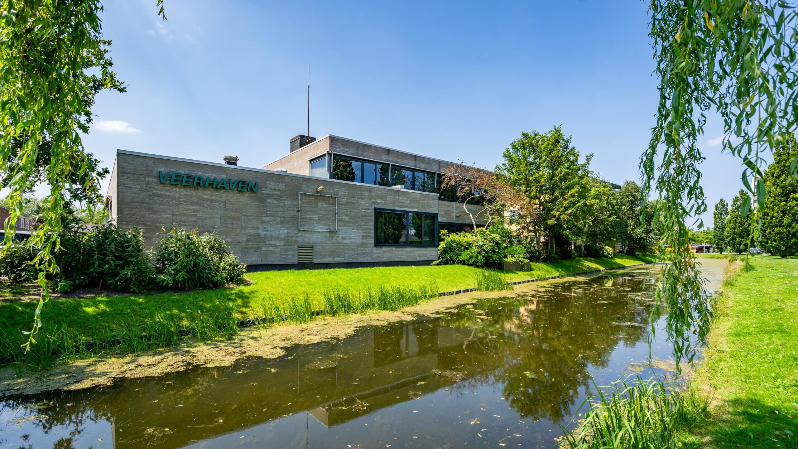 Modern gebouw met de naam "Veerhaven" naast een vijver en omgeven door groene bomen.