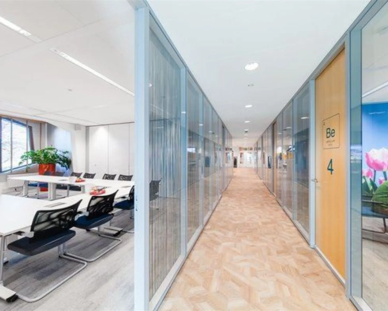 Heldere kantoorhal met glazen wanden, een vergaderruimte links en deur 4 met opschrift "Be" rechts.