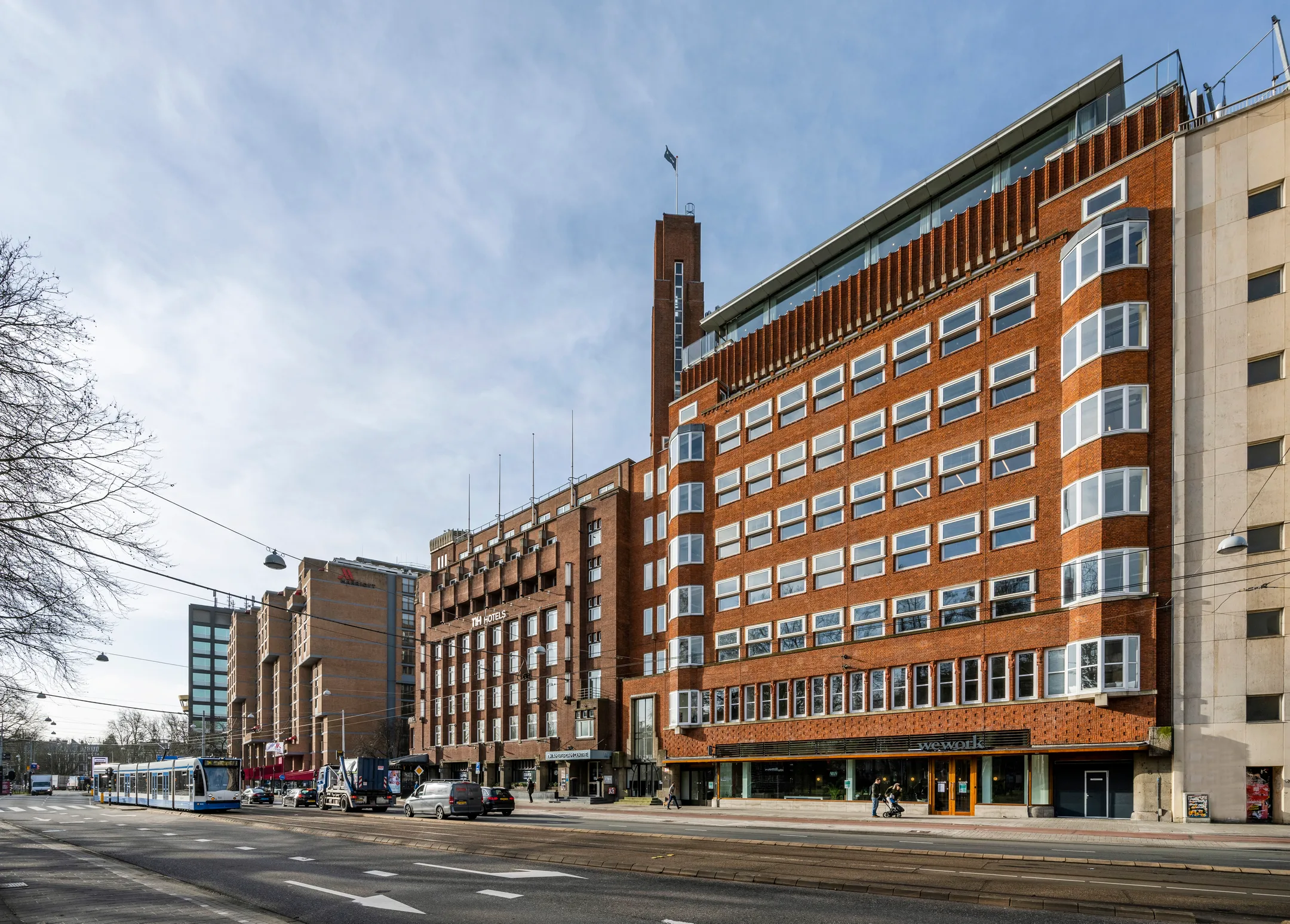 Een straatbeeld met een rij van bakstenen gebouwen, waaronder een NH Hotel, langs een weg met verkeer en een tram.