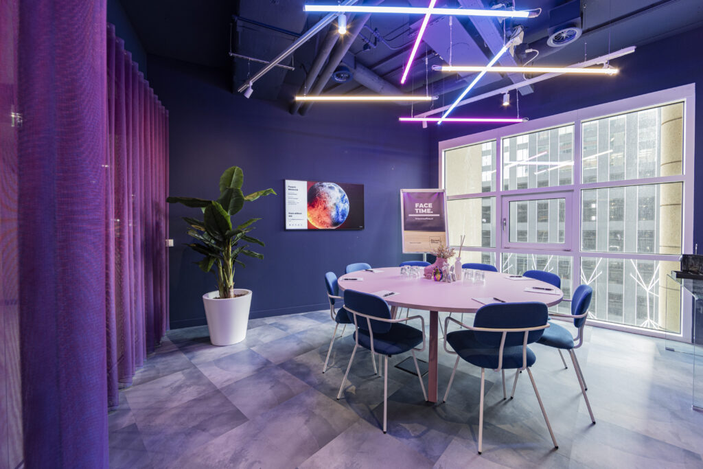 Een moderne vergaderruimte met een ronde tafel, blauwe stoelen, een groot raam en kleurrijke neonverlichting.