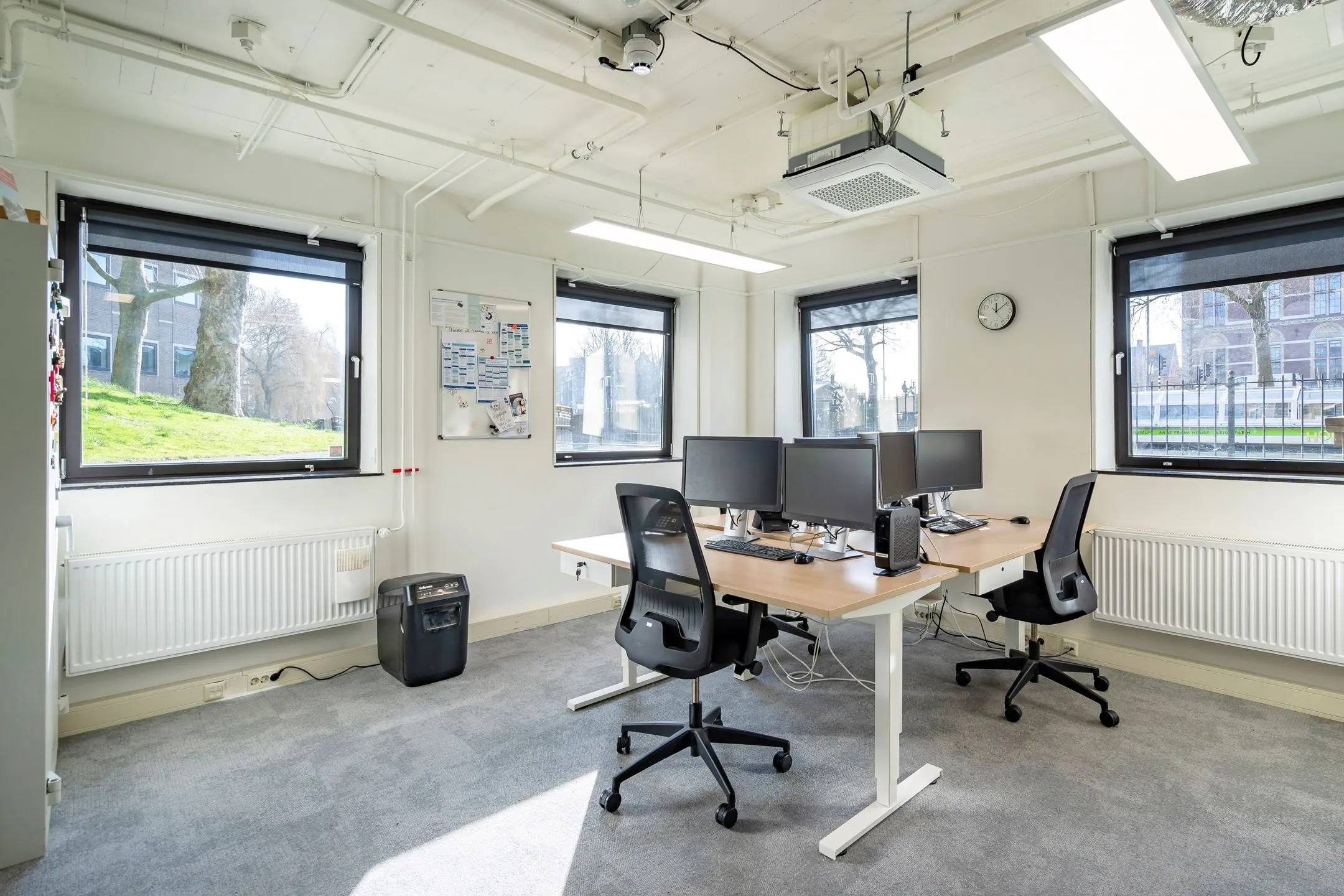 Modern, licht kantoor met vier werkplekken, bureaustoelen en meerdere computerschermen bij grote ramen.