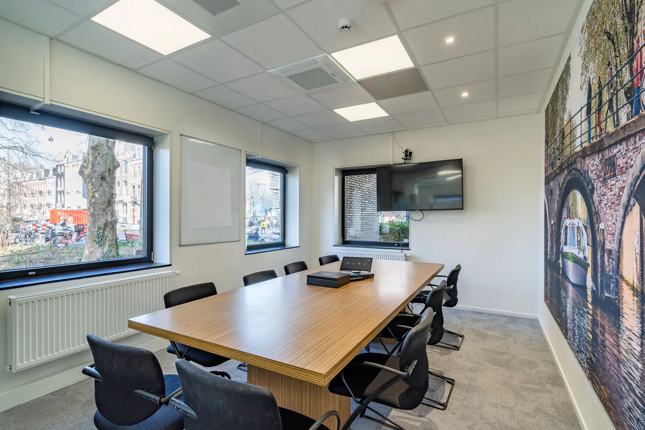 Vergaderruimte met houten tafel, zwarte stoelen, flatscreen en een grote muurprint van een Amsterdamse gracht.