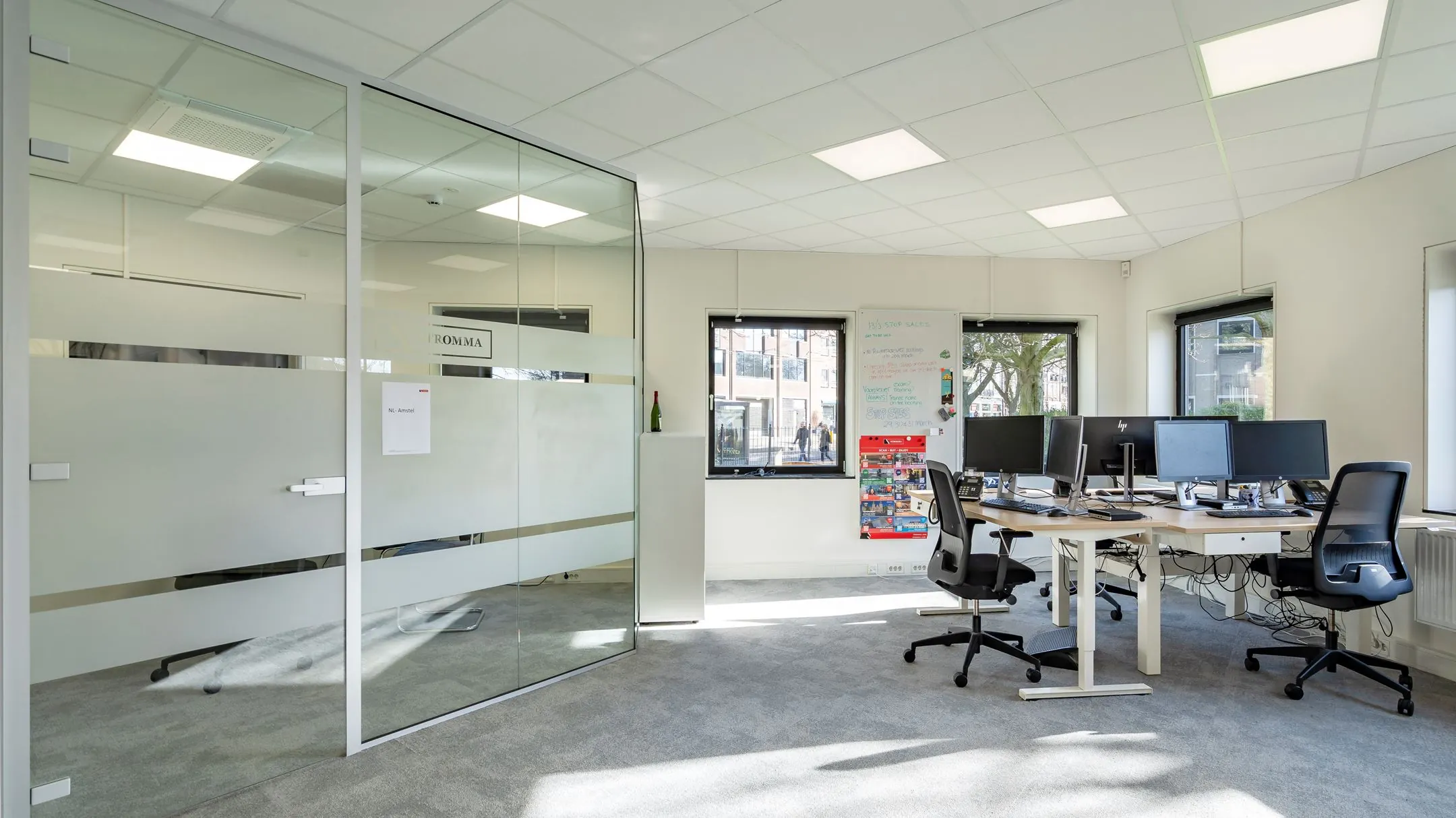 Modern kantoor met glazen vergaderruimte, meerdere bureaus met computerschermen en veel daglicht.