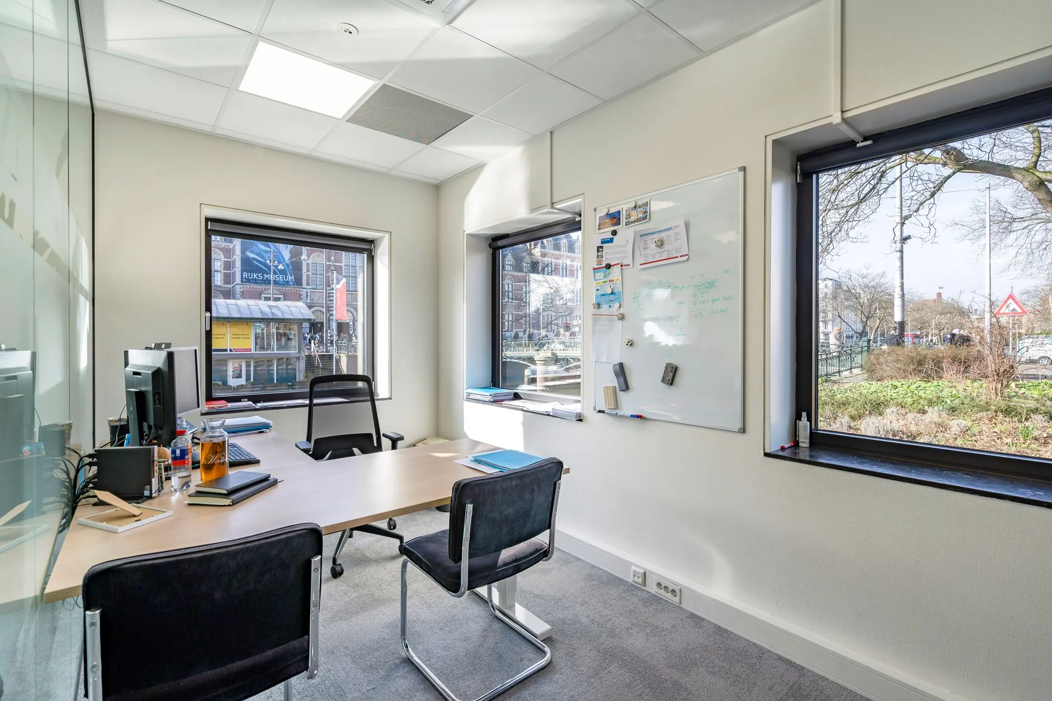 Modern kantoor met glazen wand, bureau, whiteboard en uitzicht op het Rijksmuseum in Amsterdam.