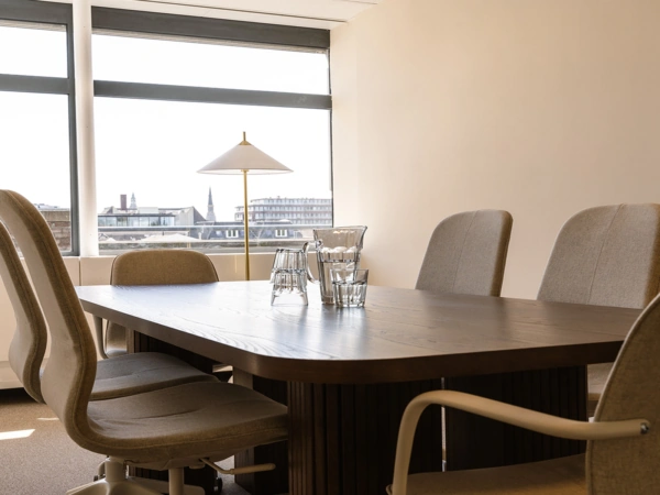 Vergaderruimte met een houten tafel, zes gestoffeerde stoelen en een karaf met glazen, met uitzicht op de stad.