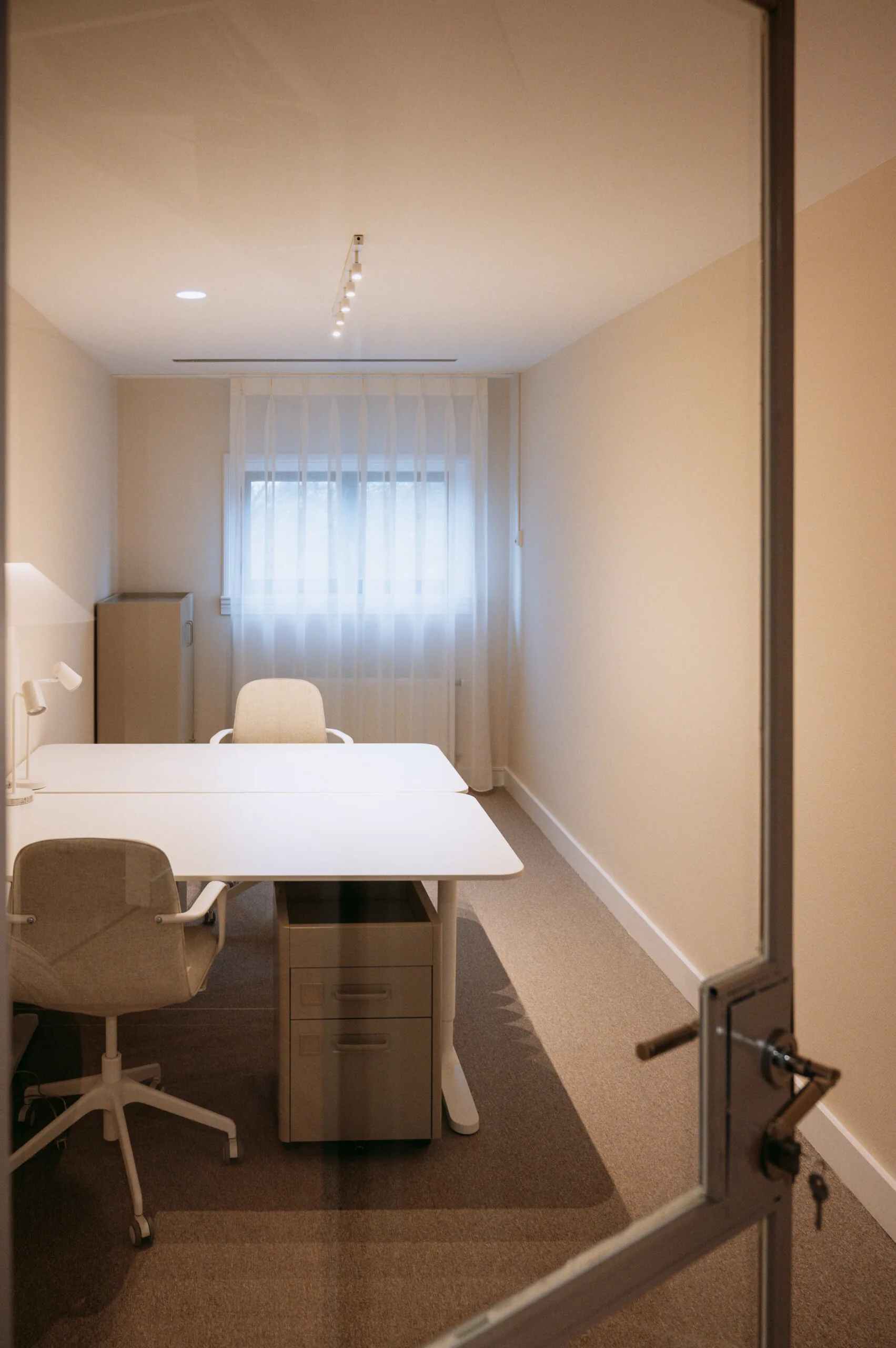 Kleine, minimalistisch ingerichte kantoorruimte met twee bureaustoelen, een wit bureau en natuurlijk licht door een raam met witte gordijnen.