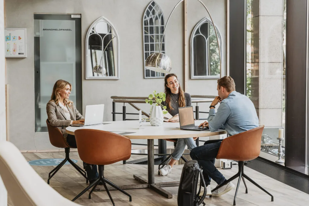 Drie mensen werken en overleggen samen aan een ronde tafel in een moderne werkruimte bij HNK Utrecht CS.