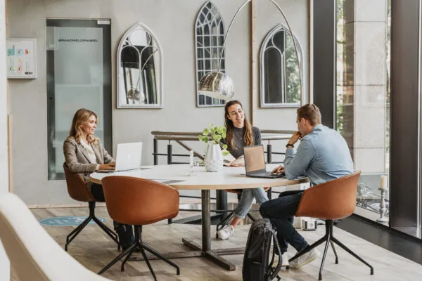 Drie mensen werken en overleggen samen aan een ronde tafel in een moderne werkruimte bij HNK Utrecht CS.