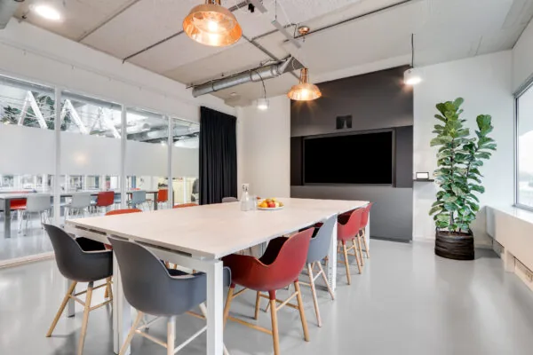 Vergaderruimte in Utrecht West met moderne inrichting, grote tafel, kleurrijke stoelen, een flatscreen en een grote kamerplant.