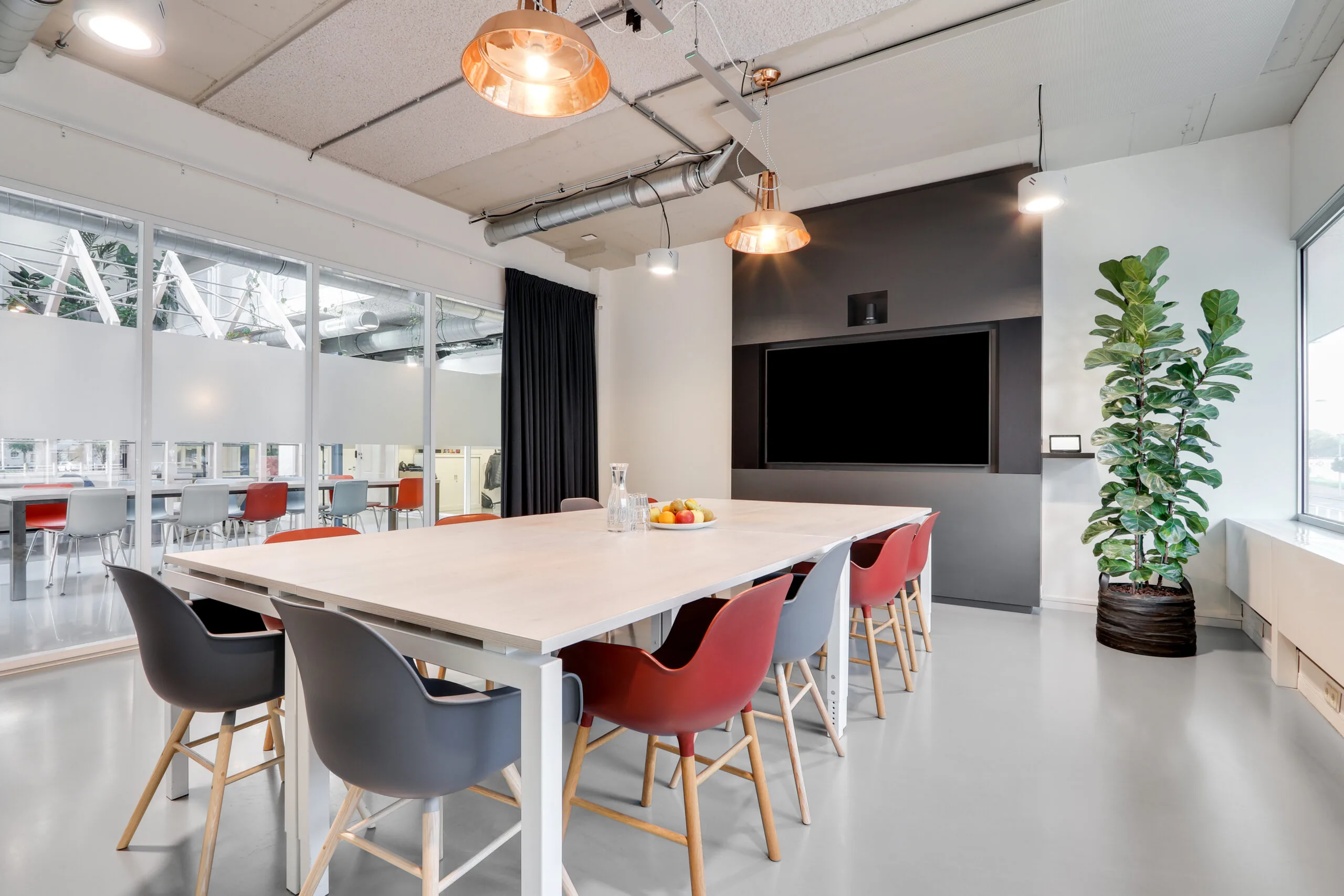 Vergaderruimte in Utrecht West met moderne inrichting, grote tafel, kleurrijke stoelen, een flatscreen en een grote kamerplant.